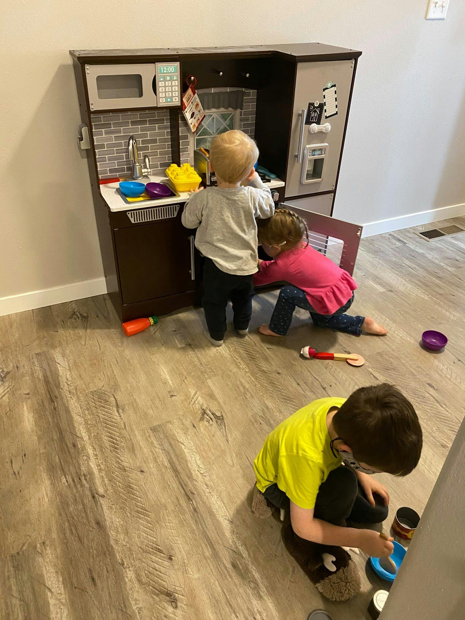 Three children playing with a toy kitchen set on a wooden floor