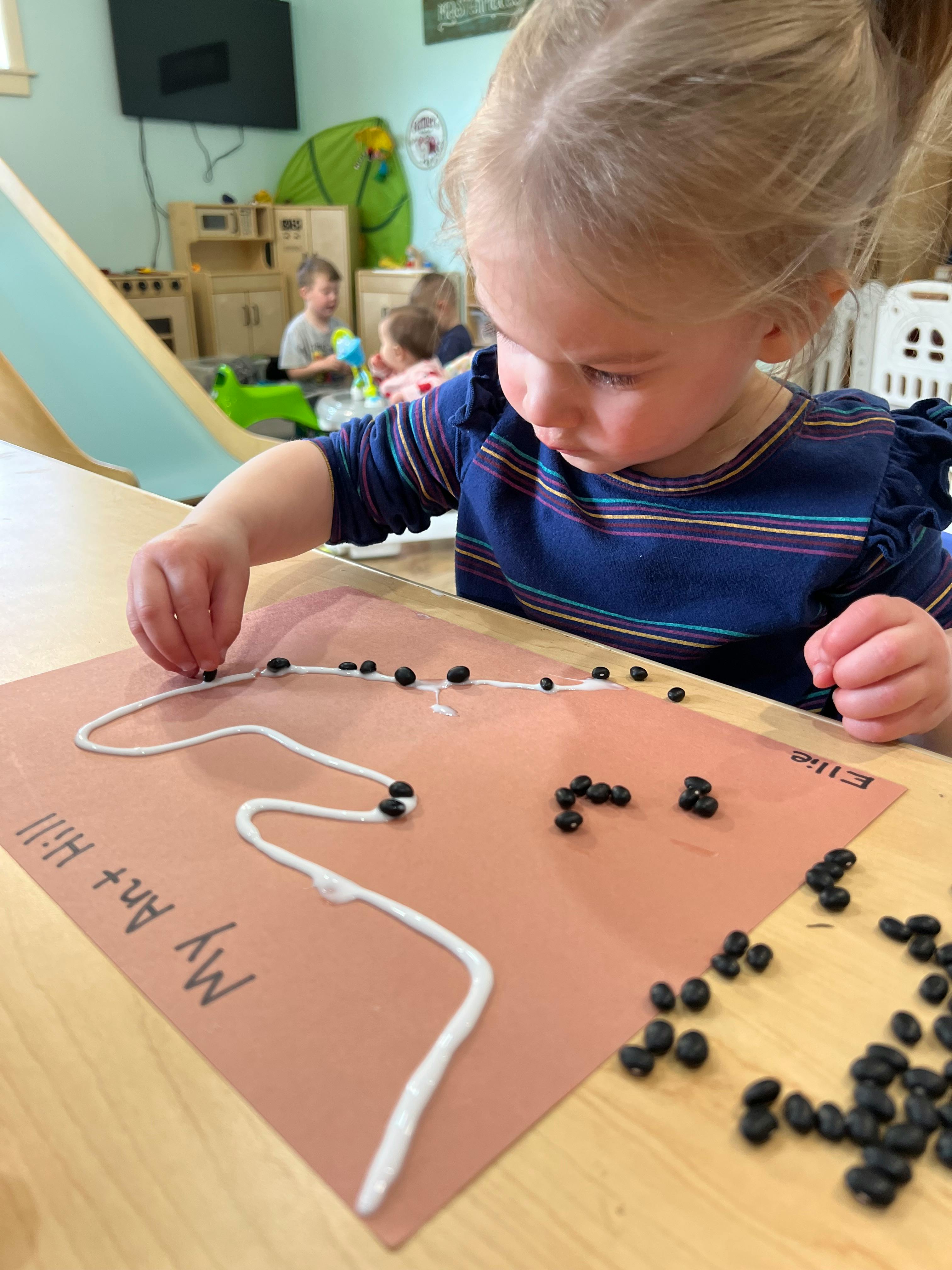 Child placing small black objects on a white string glued to paper