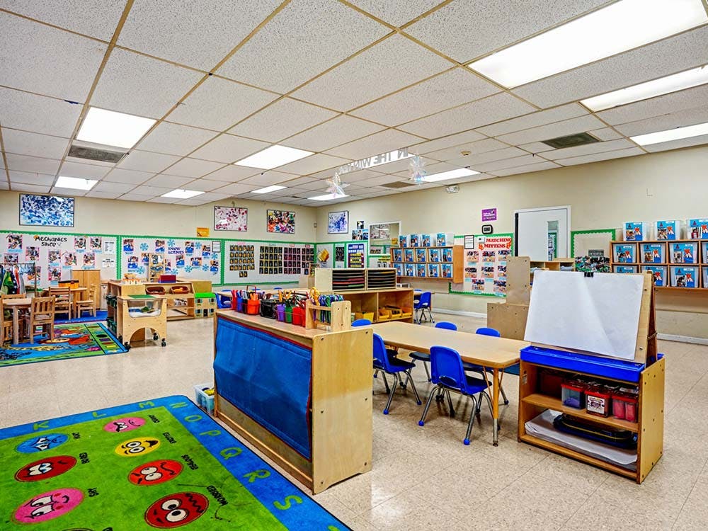 Empty classroom with small tables and chairs, colorful rugs, and learning materials