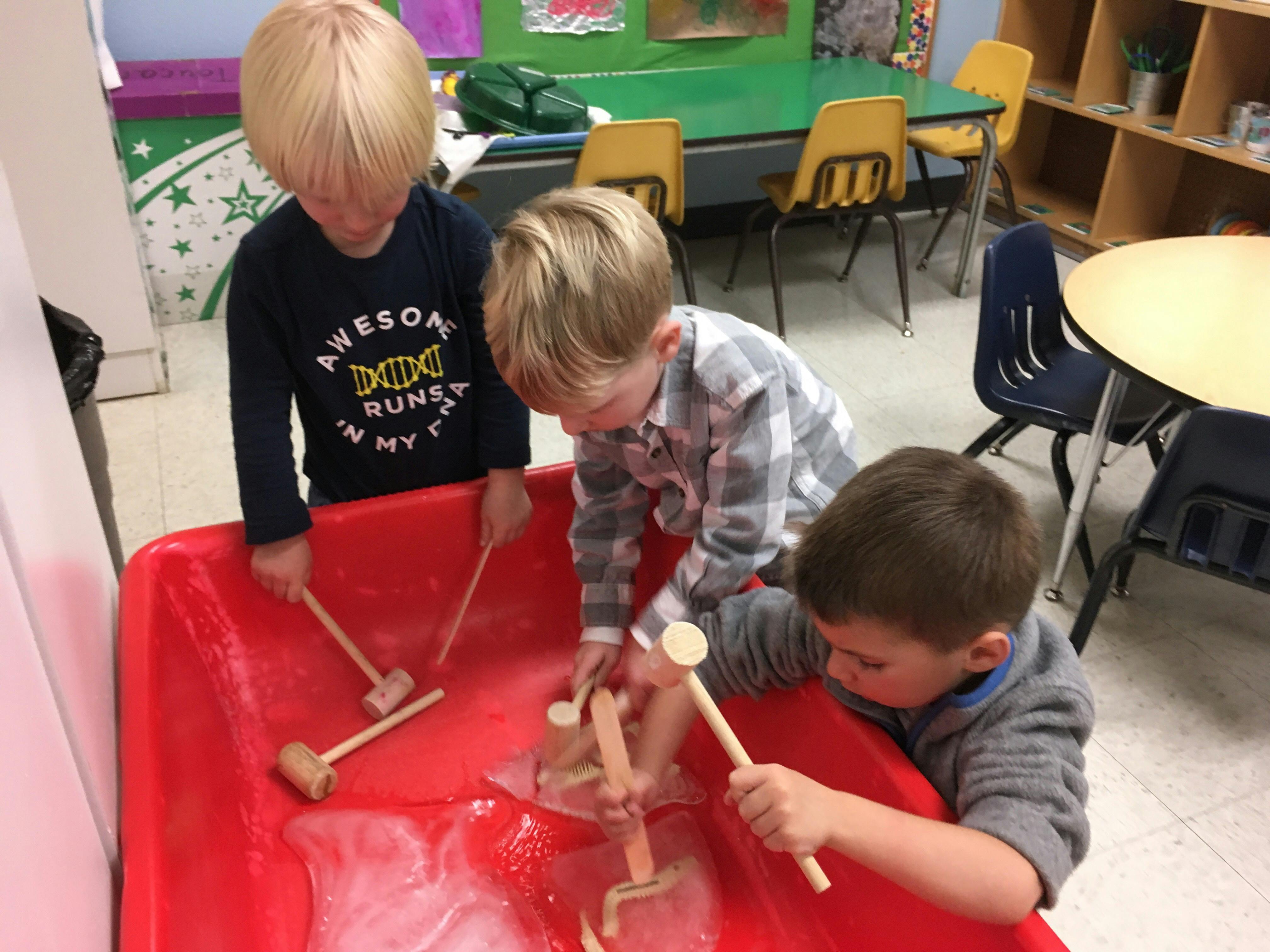 Three children playing with wooden mallets and blocks in a red bin