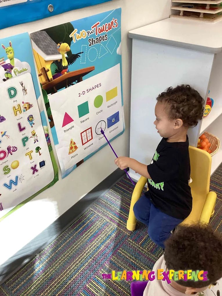Child pointing at a shapes chart with a pointer in a classroom