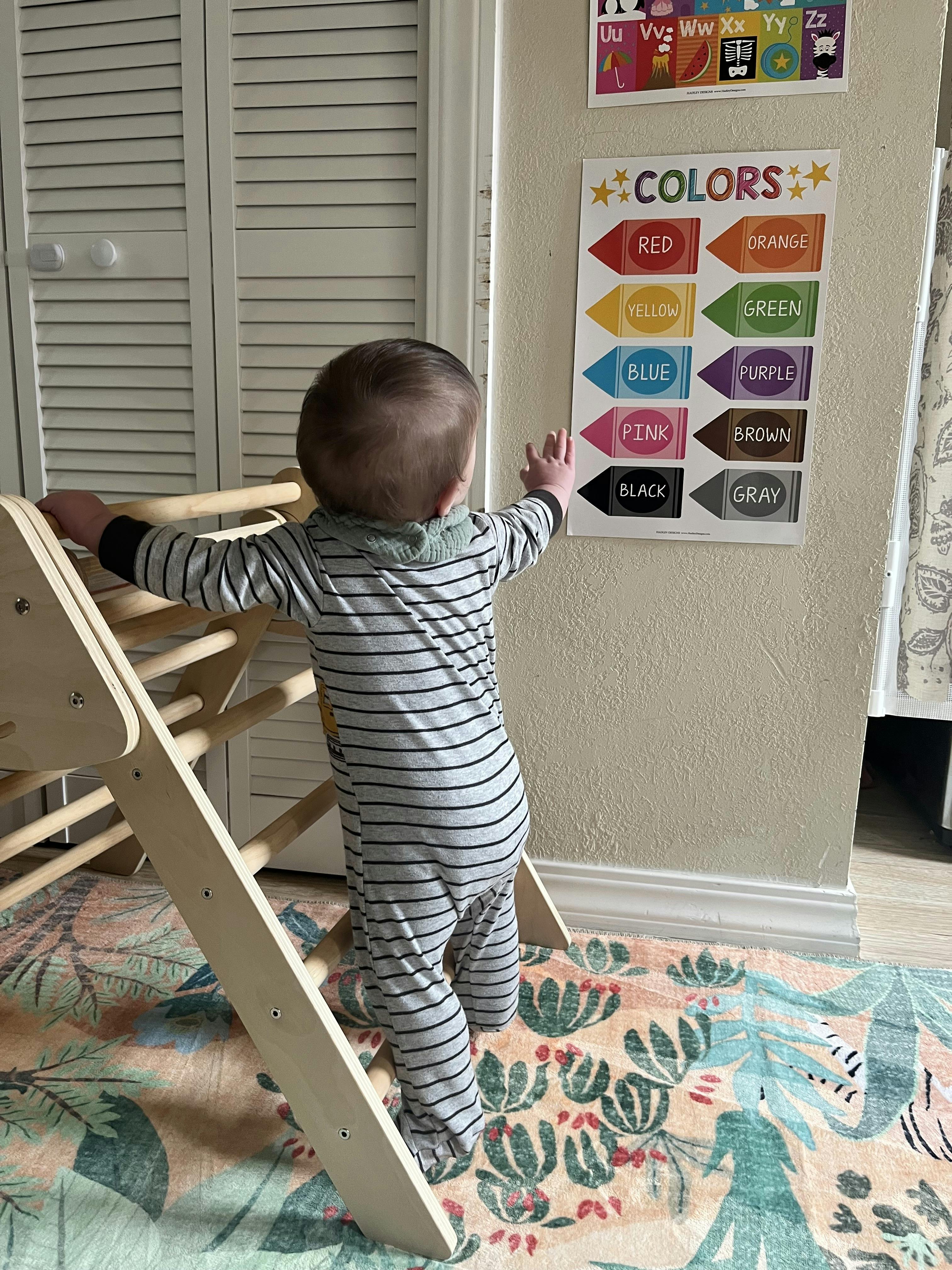 Child in striped pajamas standing on a wooden play arch, reaching for a 'COLORS' poster