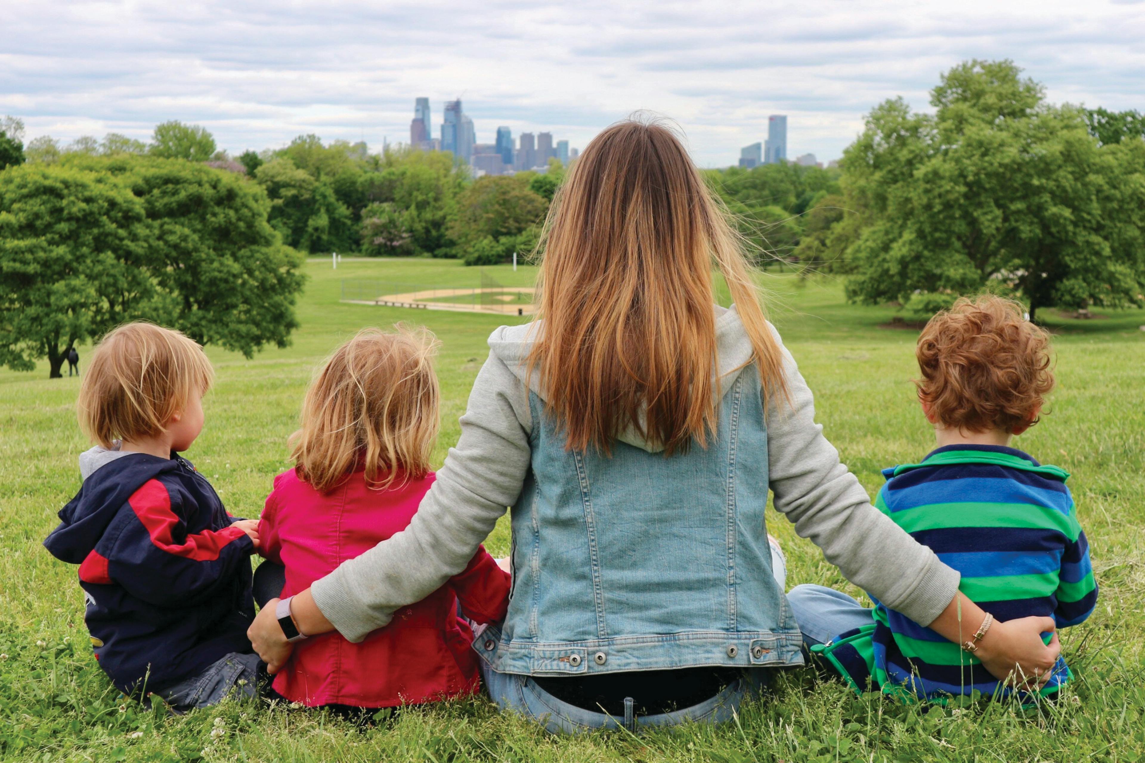 Person and three children sitting in grass looking at city skyline