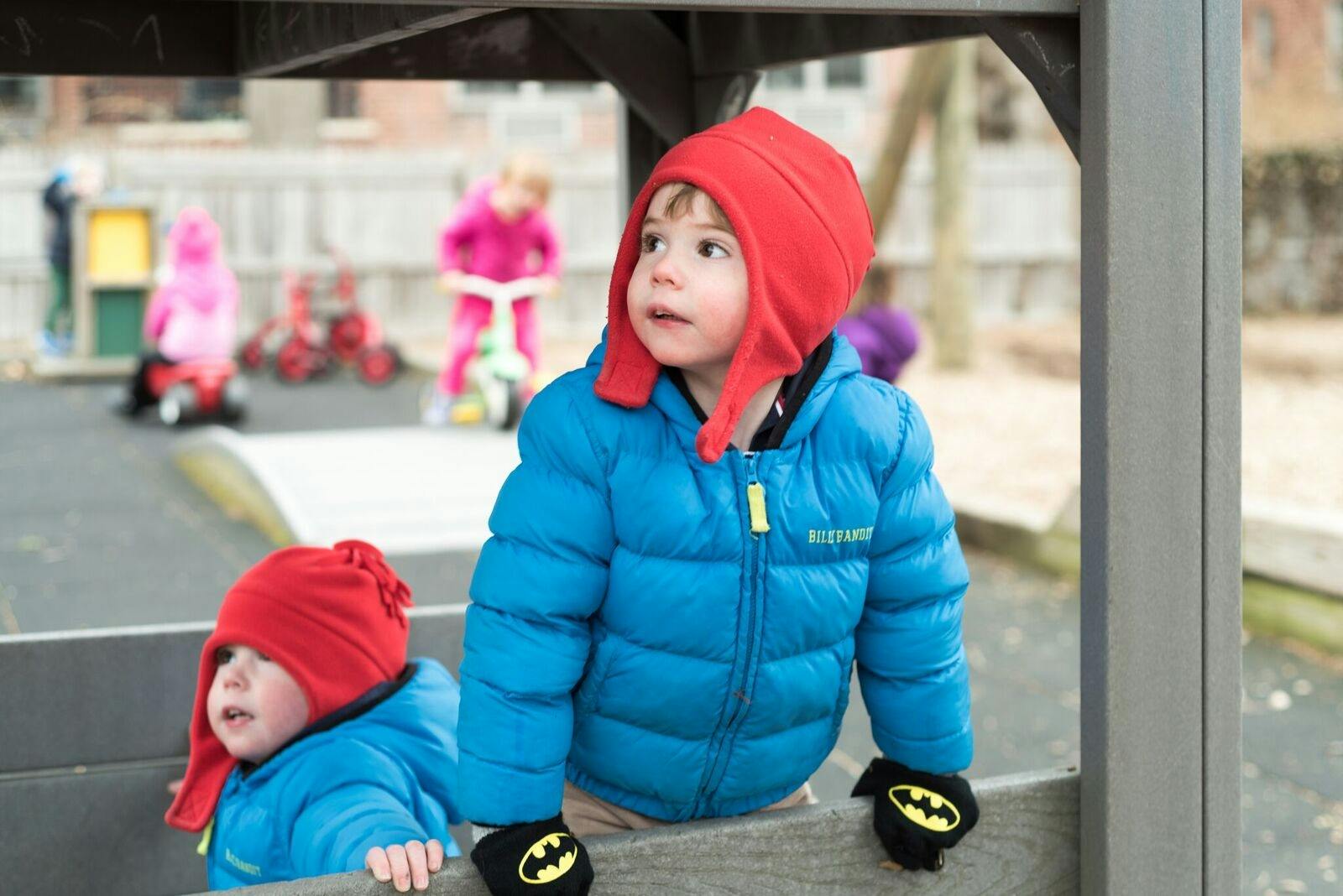 Two children in blue jackets and red hats looking out from a structure