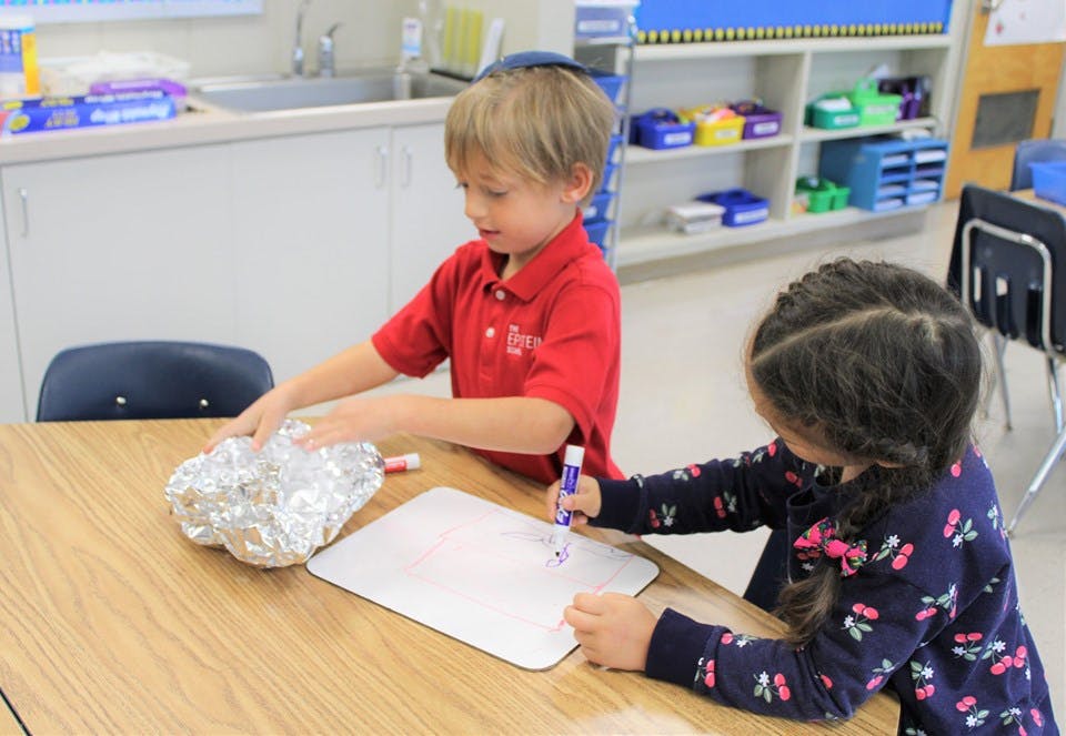 Two children at a table, one drawing and one holding a foil object