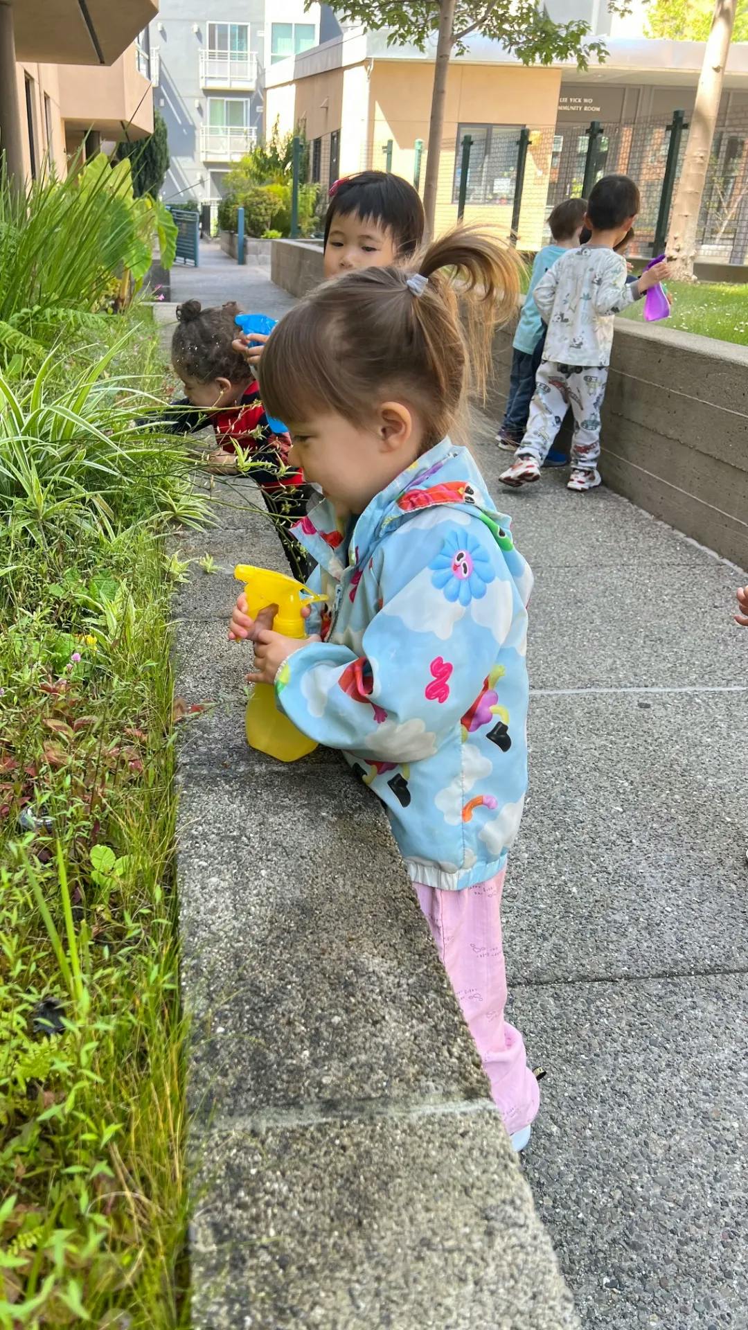 Children using spray bottles to water plants along a stone ledge