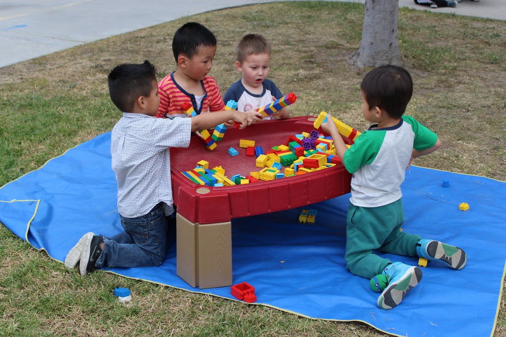 Four children playing with building blocks at a red table outdoors