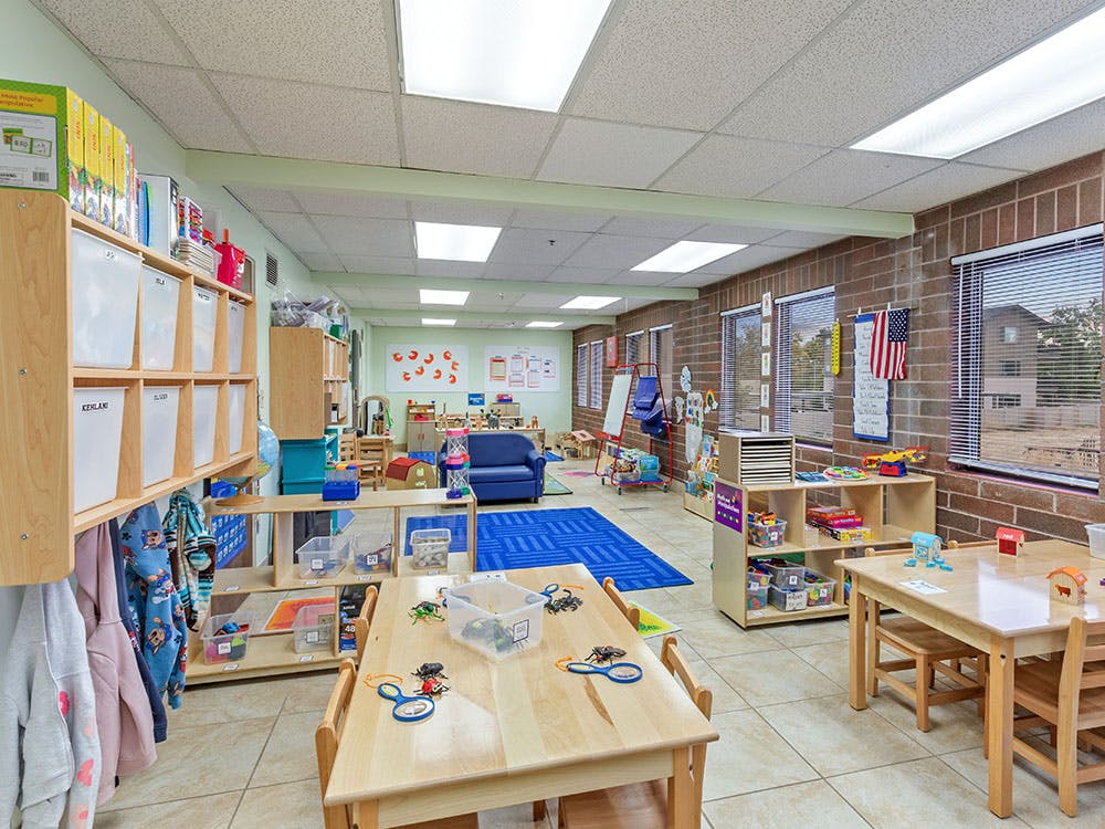 Classroom with small tables and chairs, shelves with toys, and blue rug