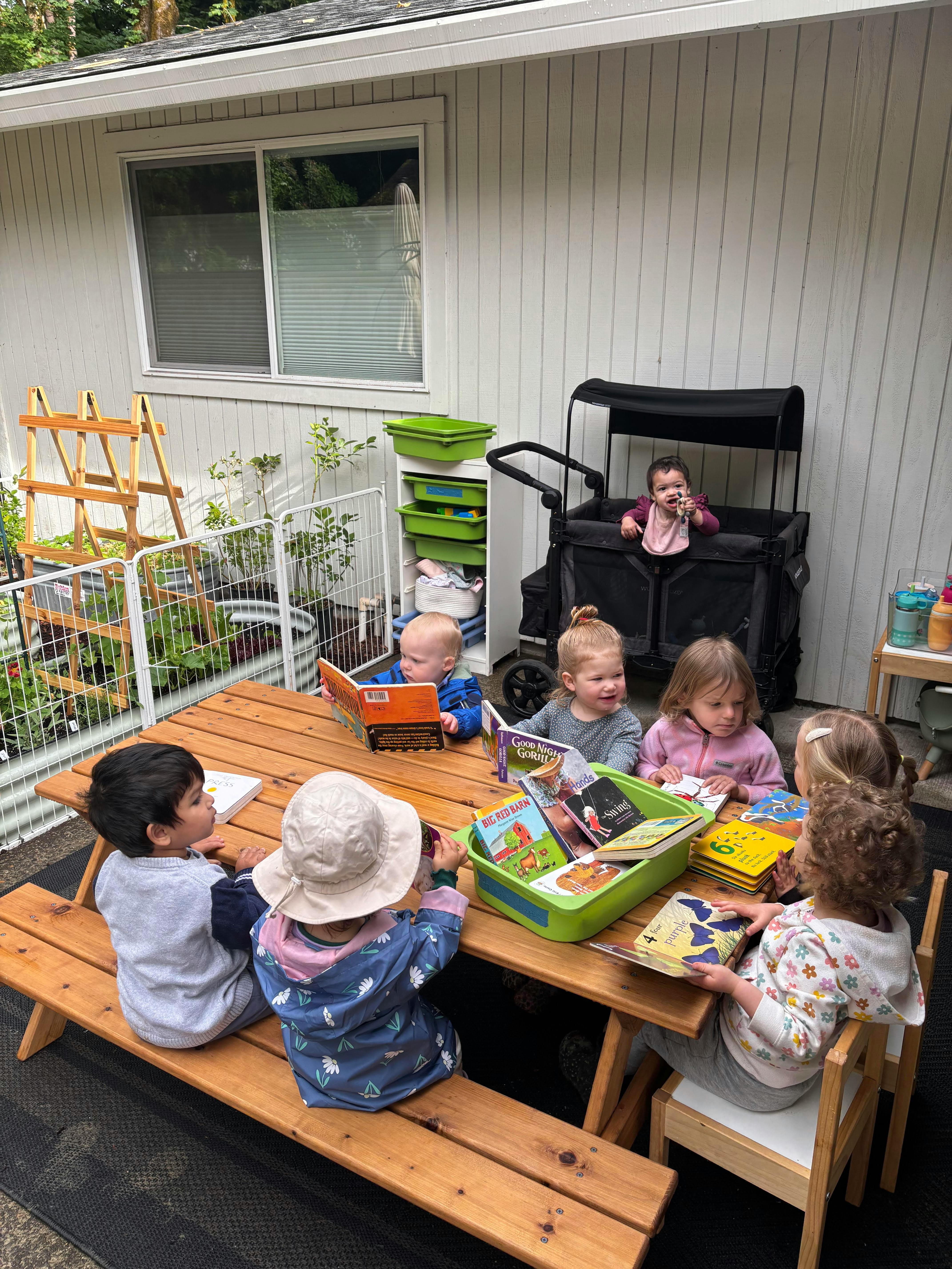 Group of children sitting at a wooden table outdoors reading picture books