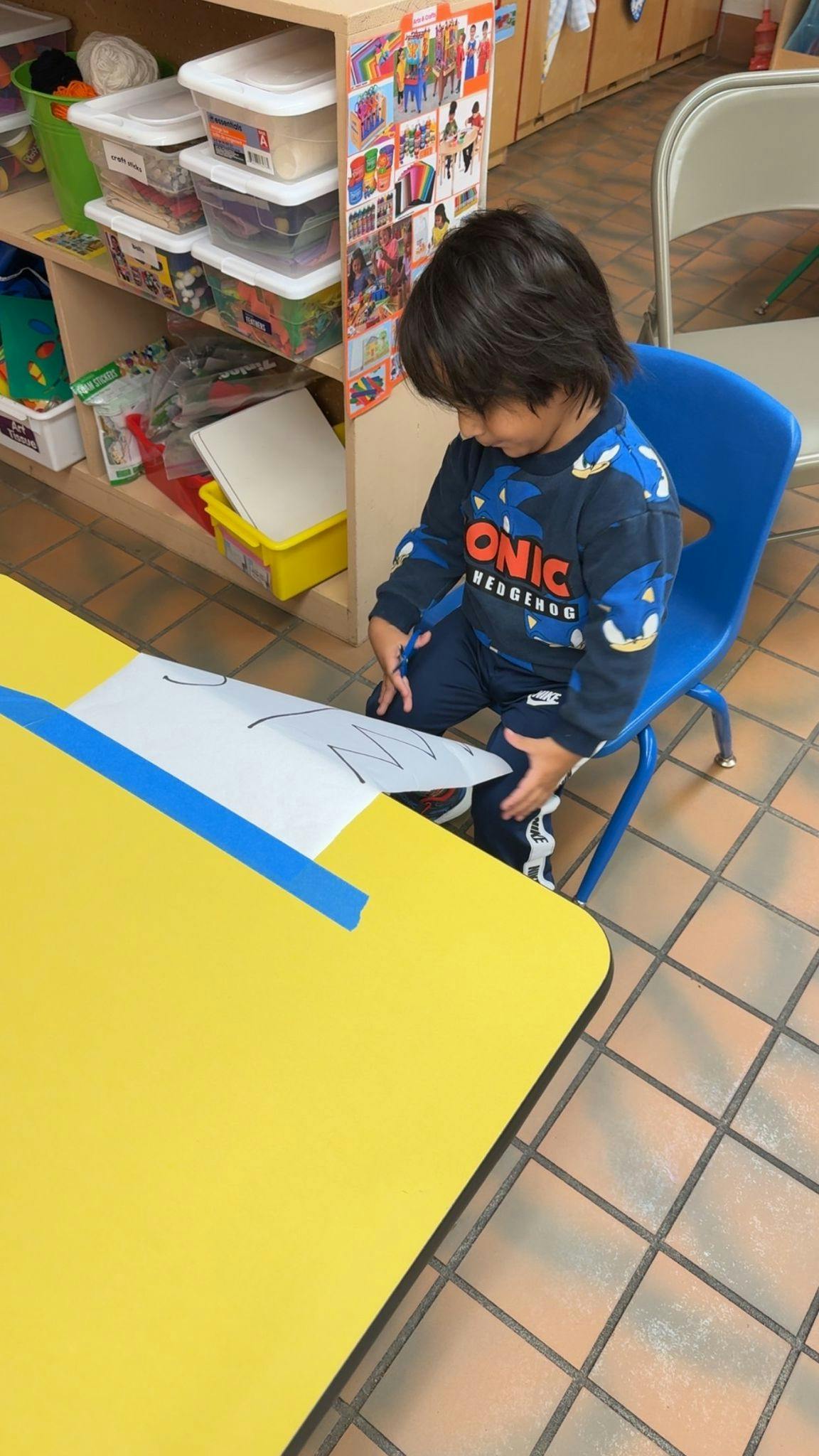 Child sitting at a yellow table cutting paper with scissors