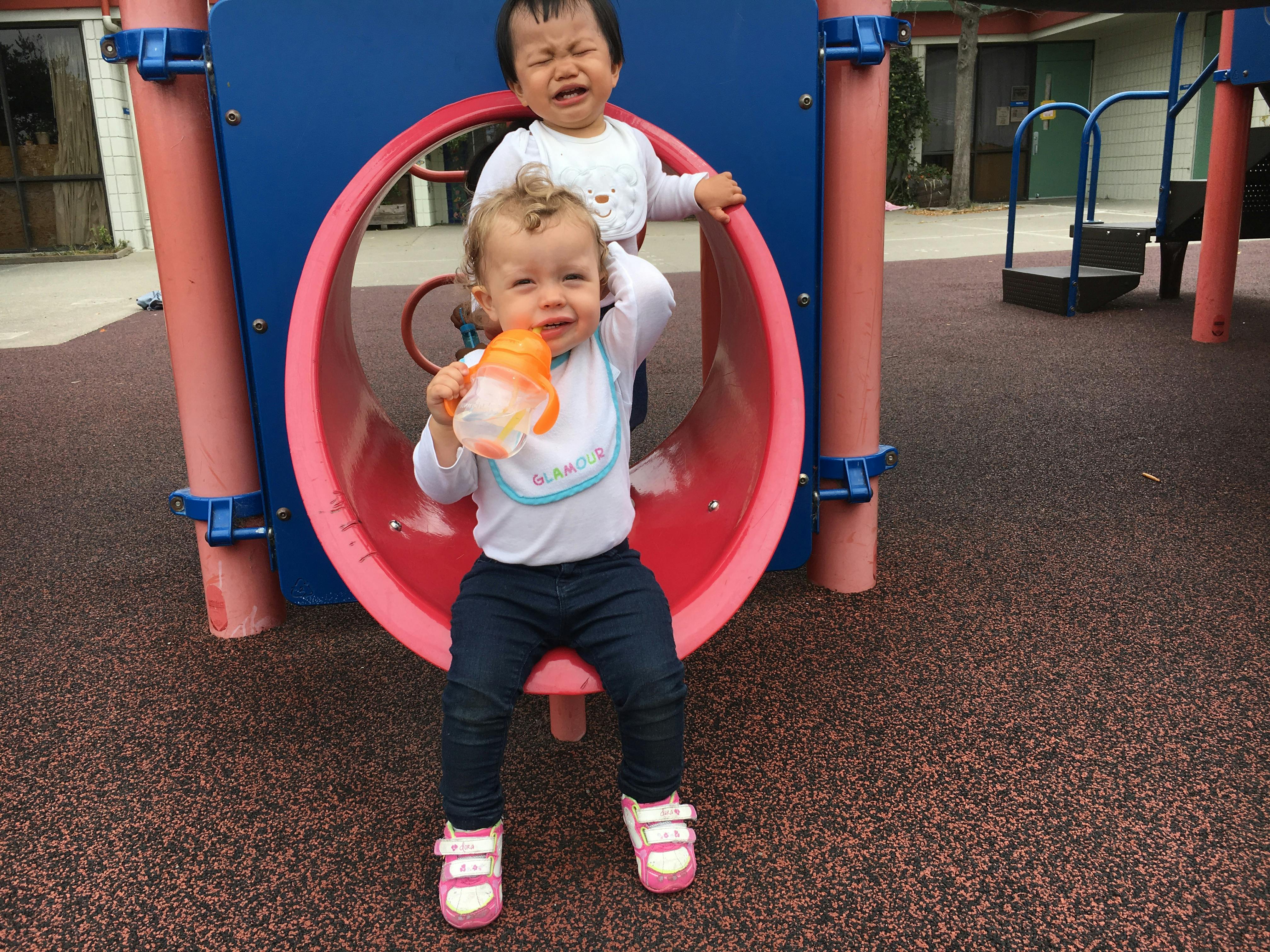 Child sitting on playground equipment with another child standing behind crying
