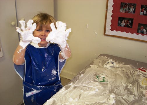 Child in a blue smock with hands covered in white foam