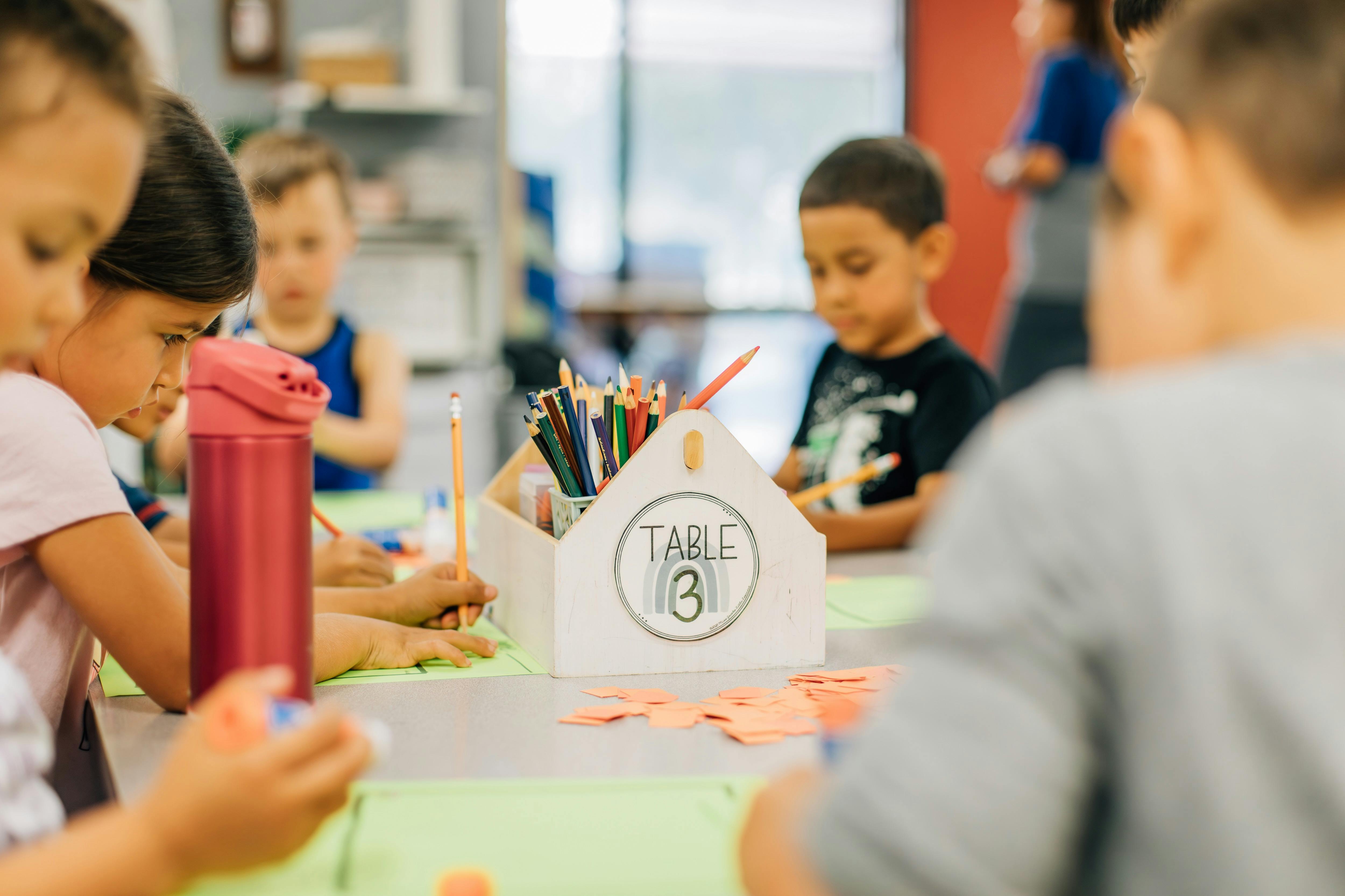 Children drawing at a table with a pencil holder labeled "Table 3"