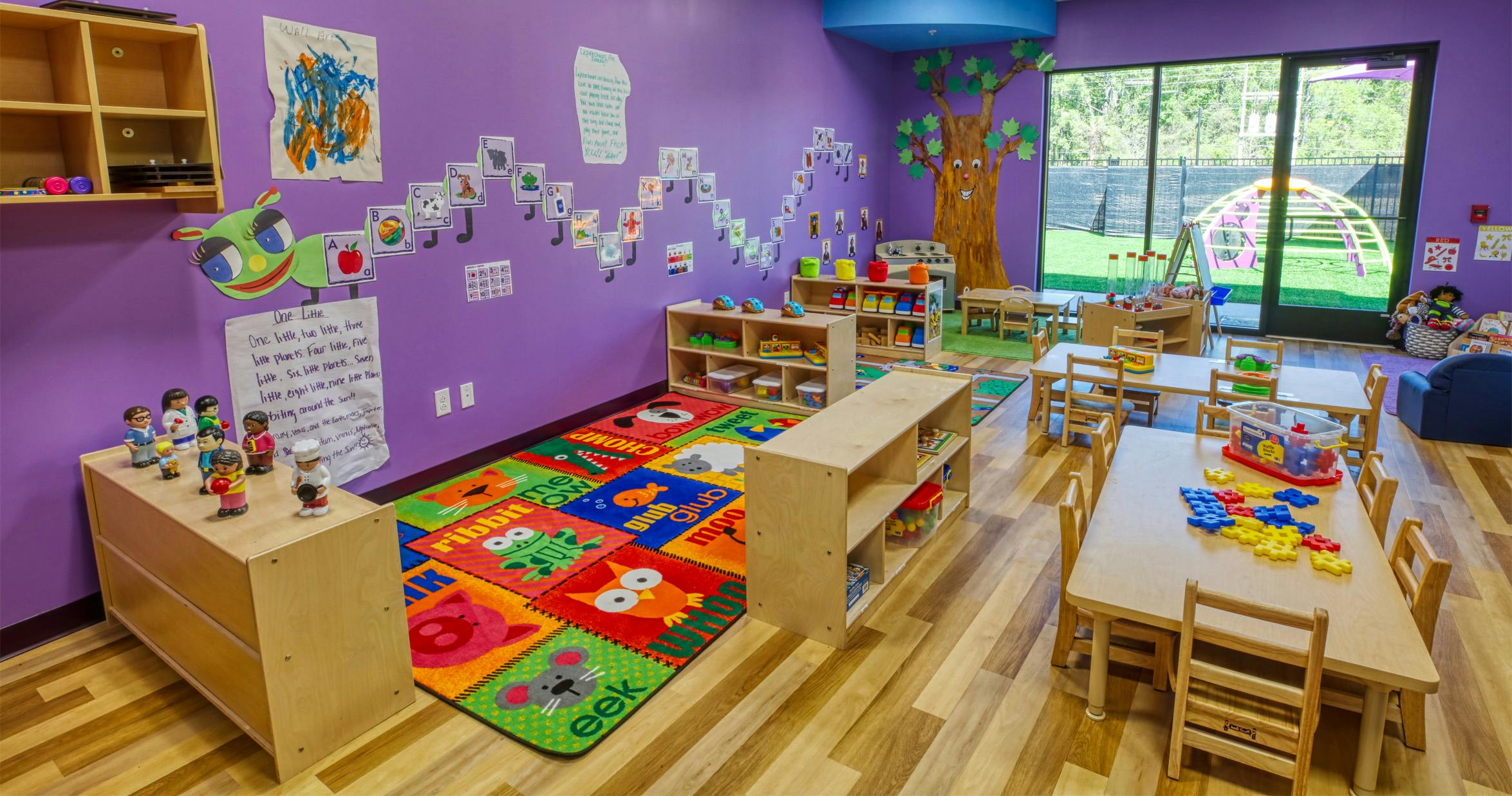 Colorful preschool classroom with tables, chairs, shelves, and a patterned rug