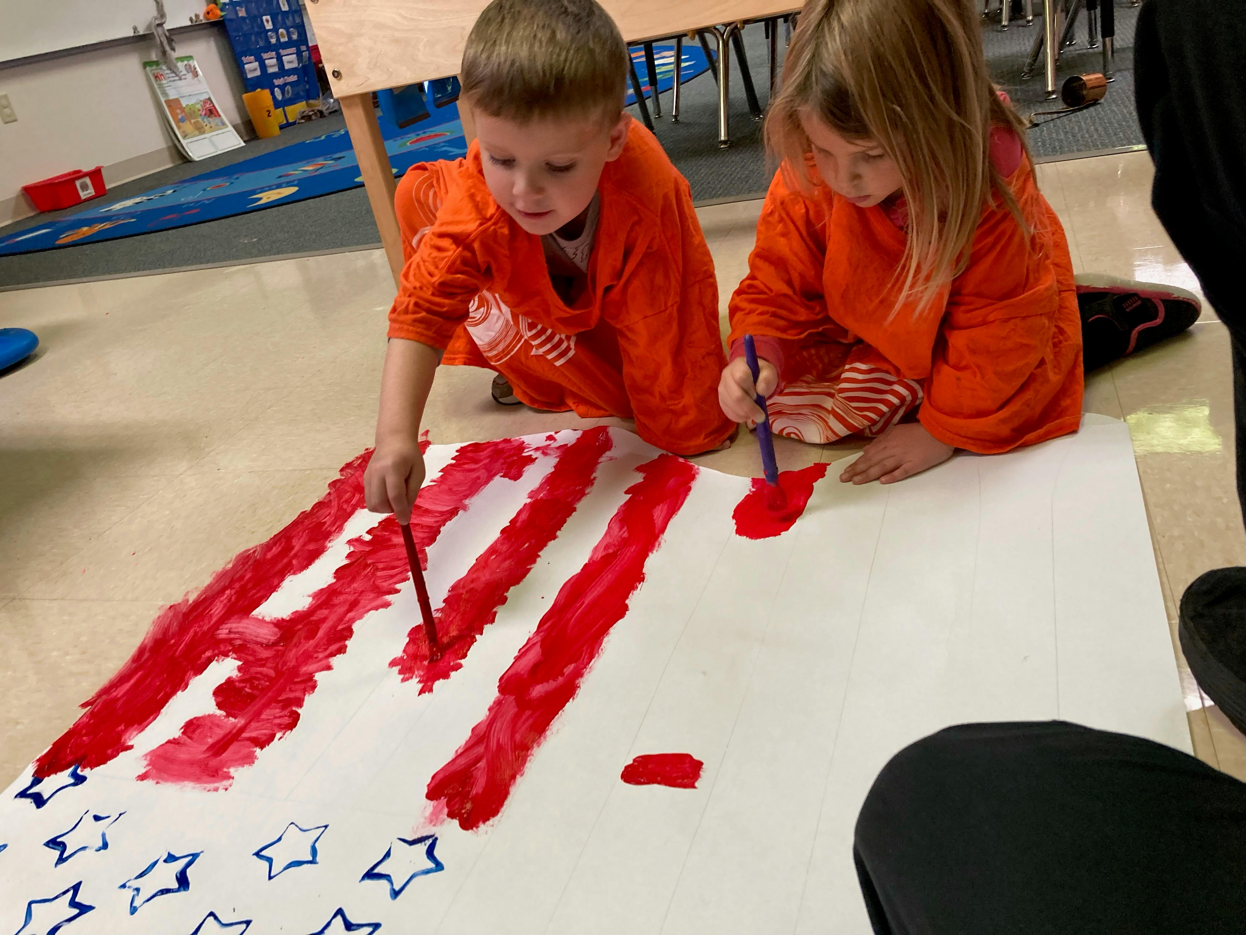 Two children painting red stripes and blue stars on a large white sheet