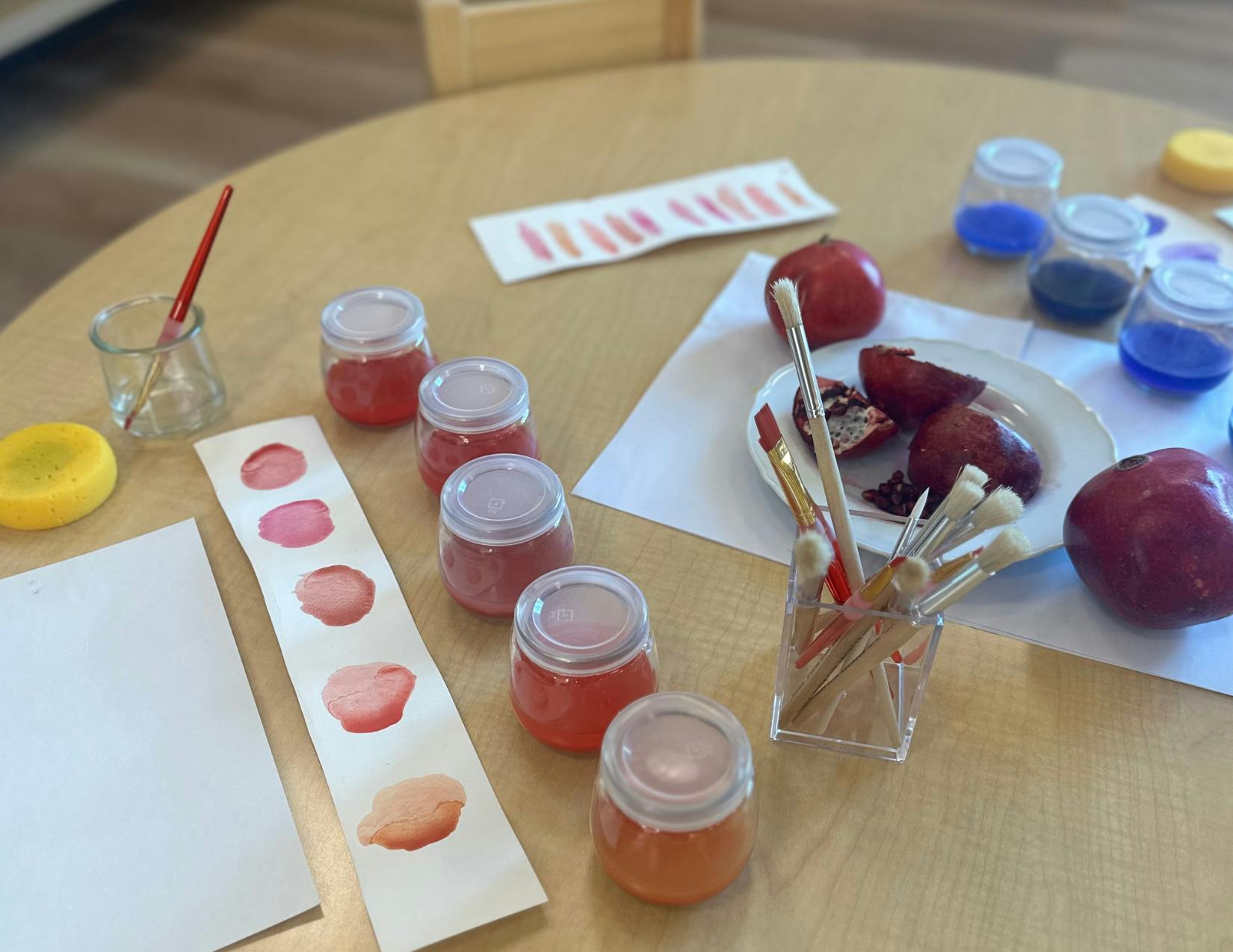 Art supplies on a table with paint jars, paintbrushes, and pomegranates