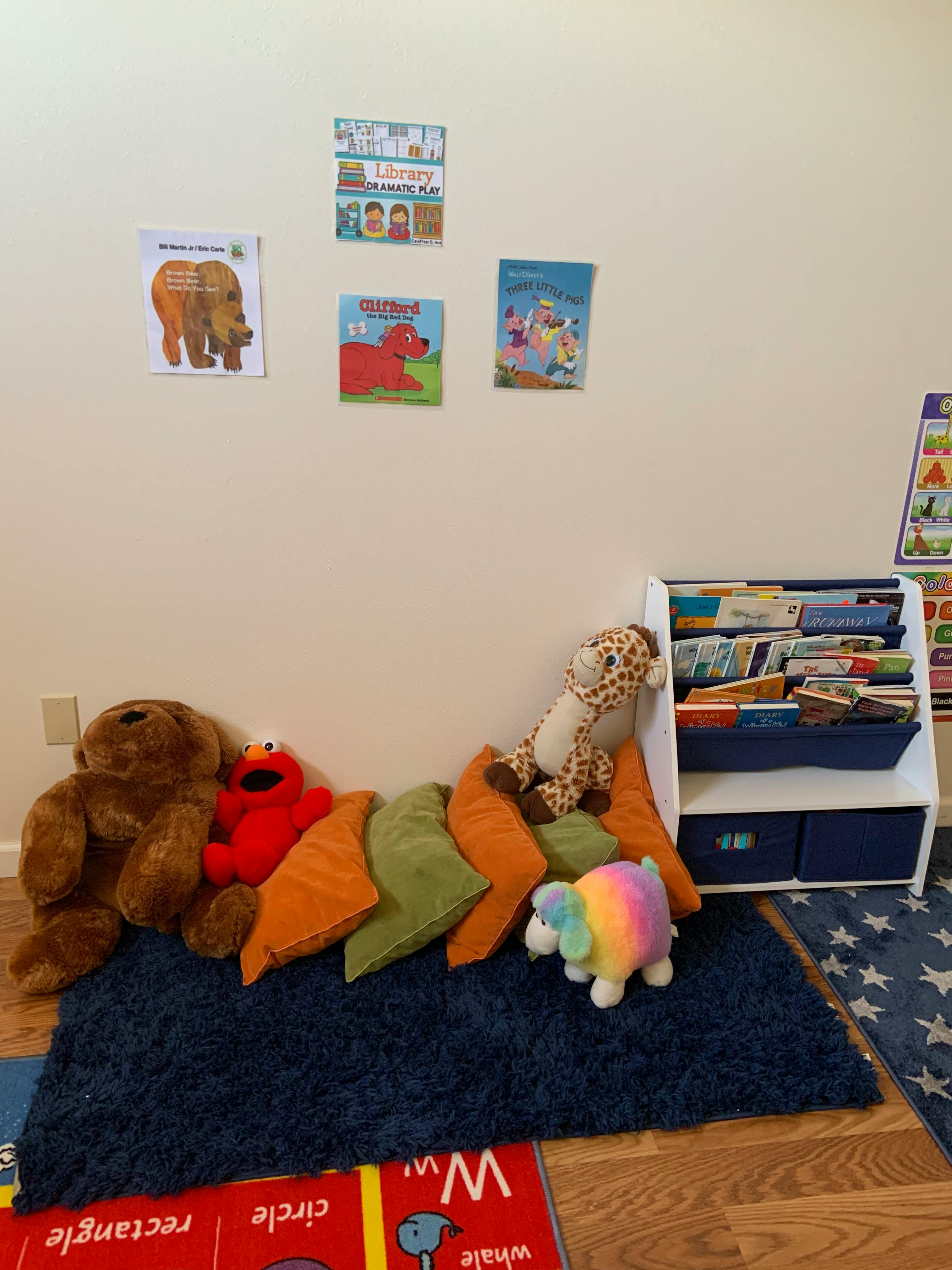 Stuffed animals and pillows on a rug in a classroom with books on the wall