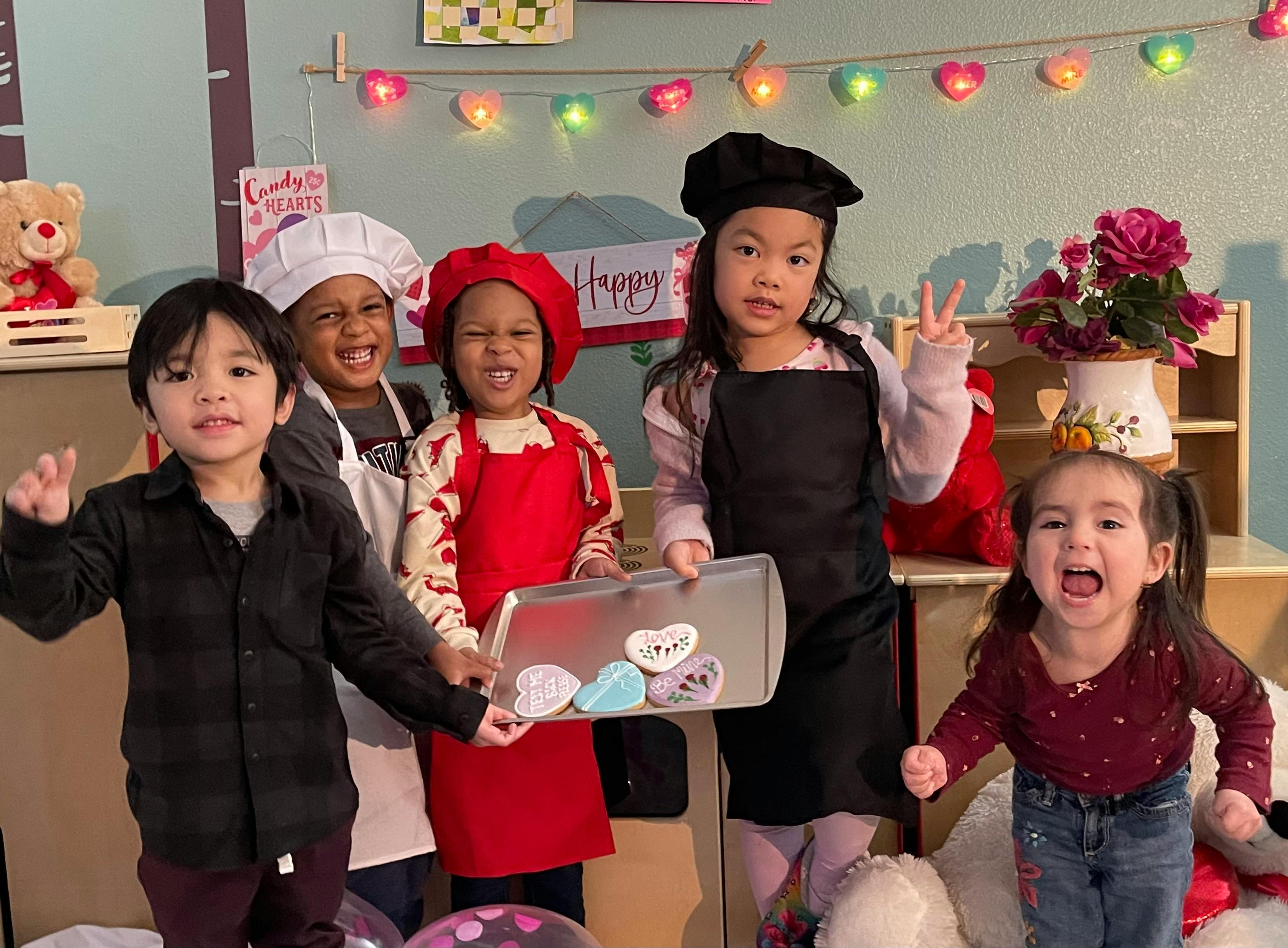 Children in chef hats and aprons holding a tray of heart-shaped cookies