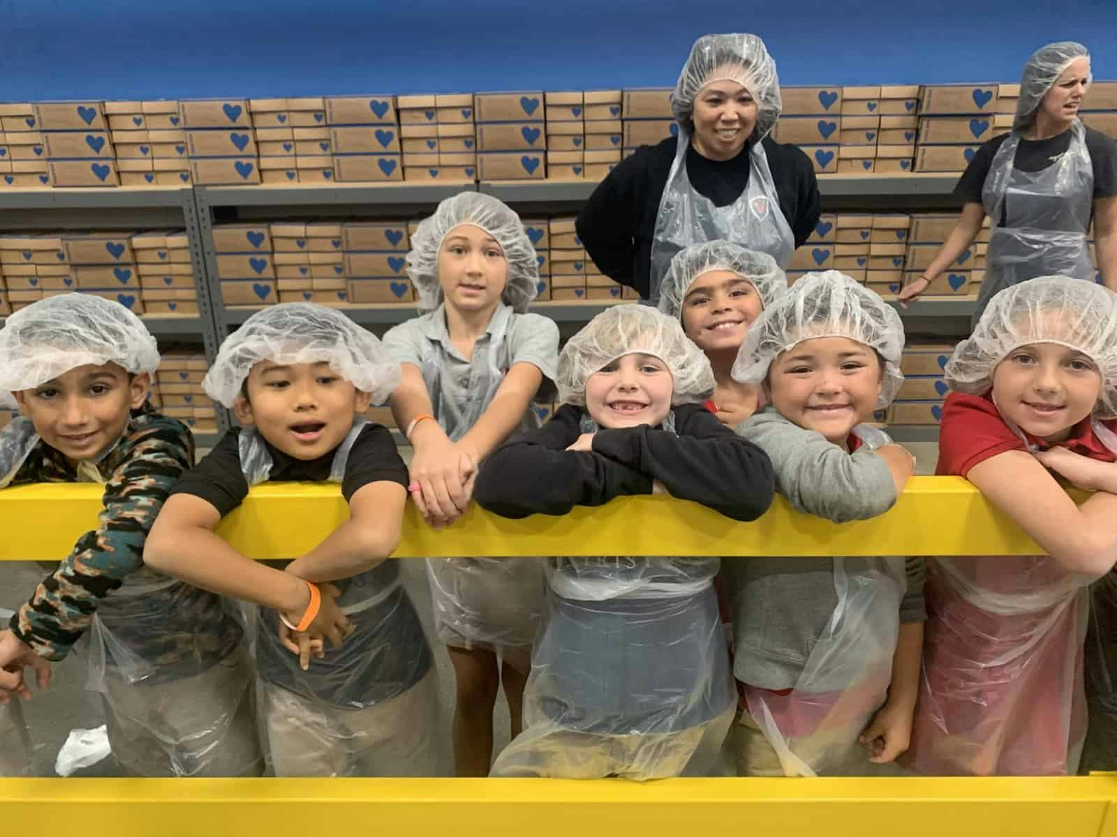 Children and adults wearing hairnets and aprons in a room with shelves of boxes