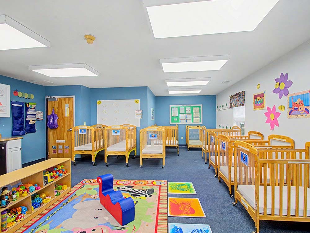 Room with multiple wooden cribs, a play rug, and shelves with toys