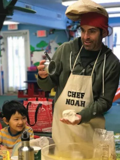 Person in "Chef Noah" apron holding dough and measuring cup, child watching