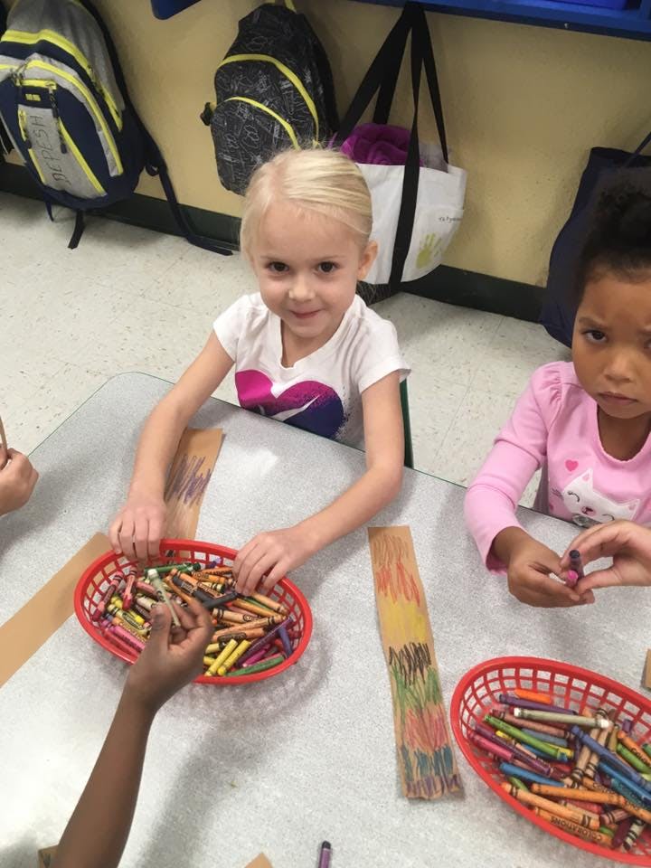 Child at a table with crayons, and reaching for, a basket of crayons