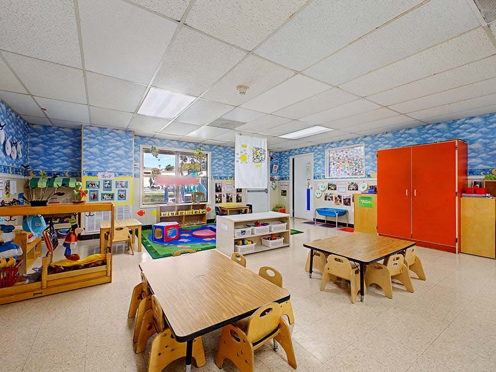 Classroom with small tables and chairs, toys, and bright blue cloud-patterned walls