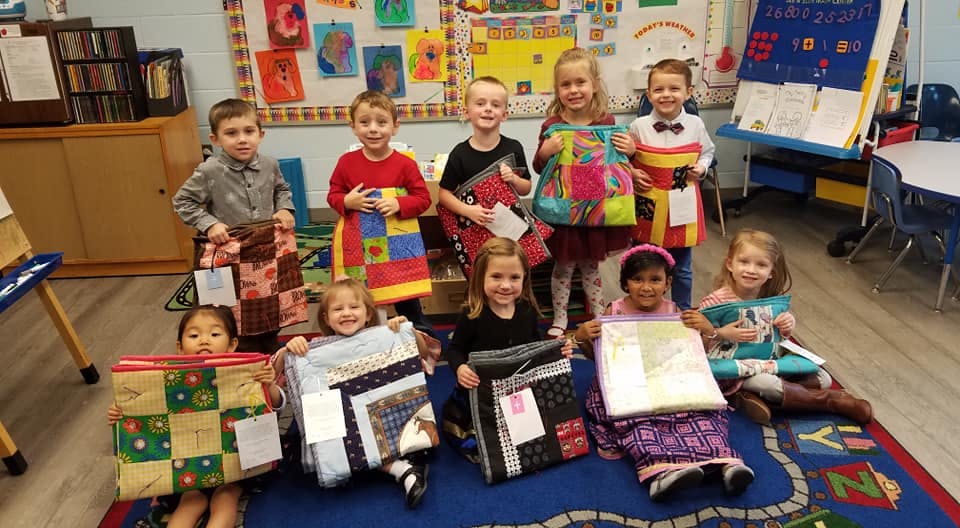 Group of children holding handmade quilts in a classroom