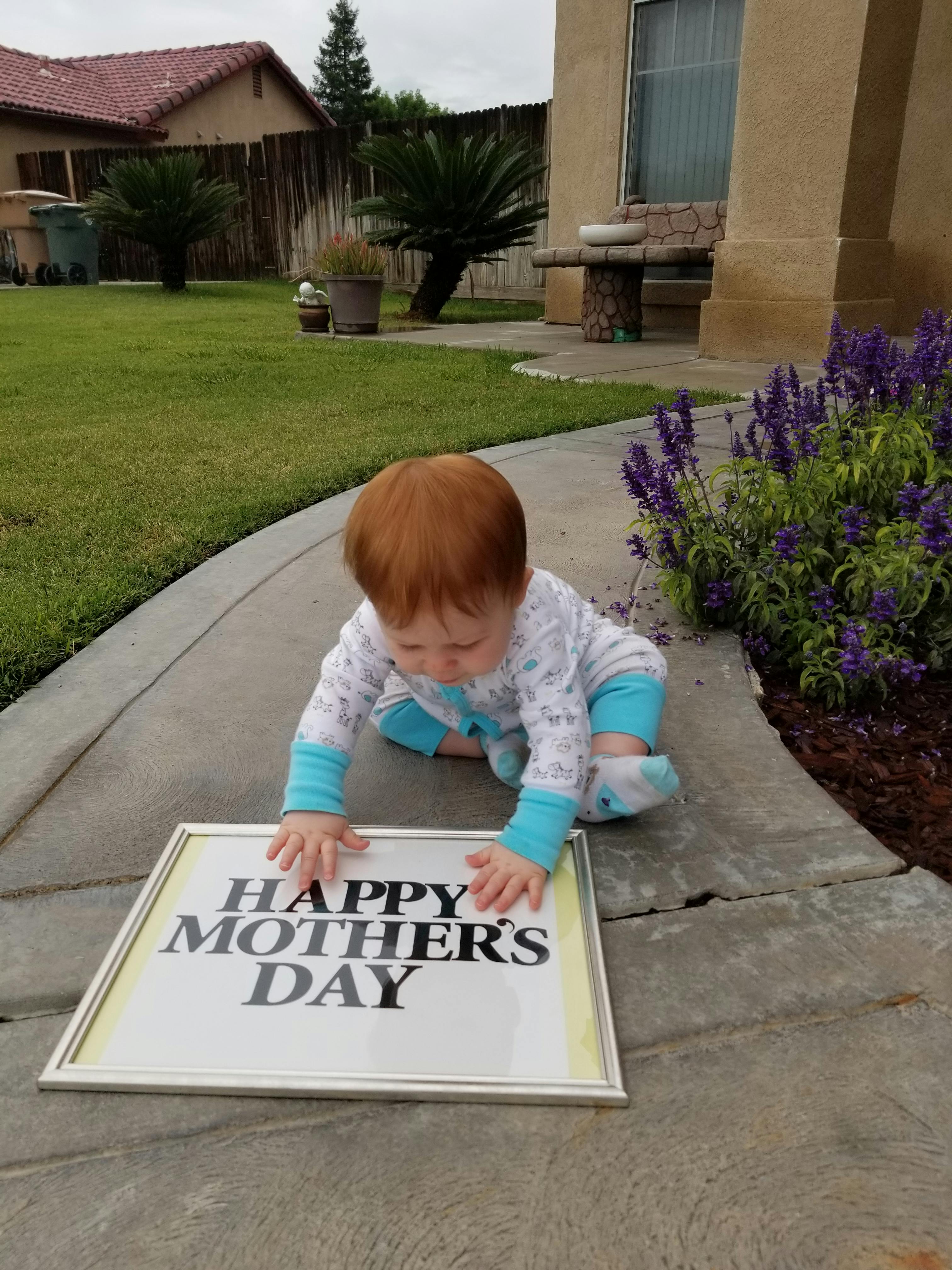 Child crawling on a sidewalk, touching a framed sign that reads "HAPPY MOTHER'S DAY"