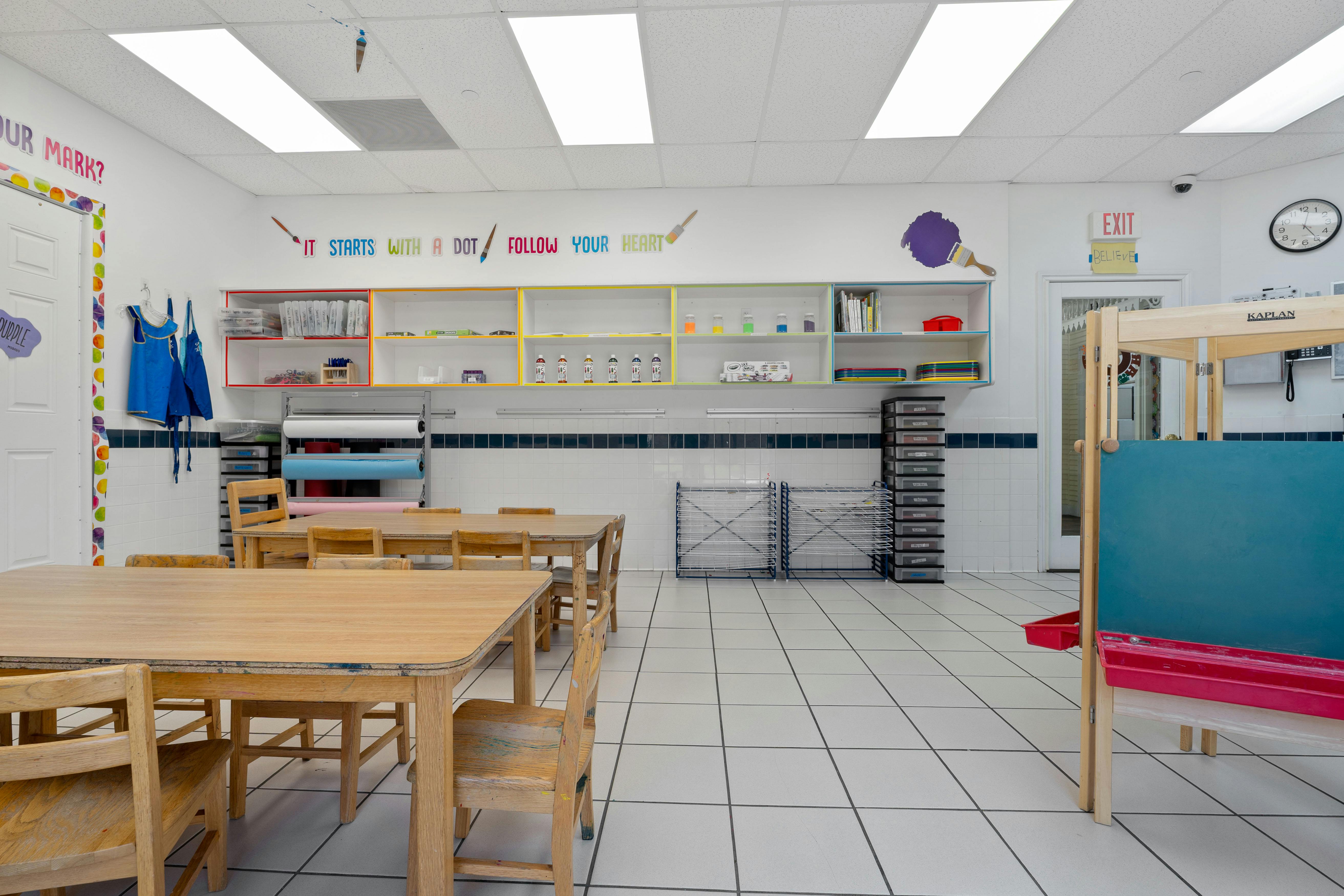 An empty classroom with several wooden tables and chairs, art supplies
