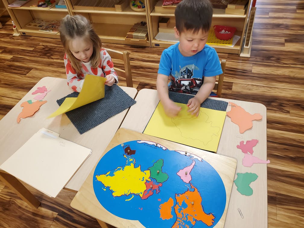 Two children at tables with a world map puzzle and craft paper