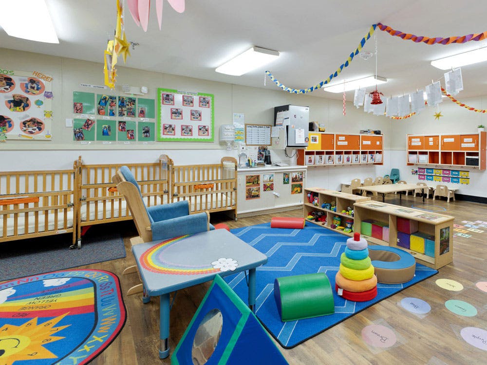 A childcare room with cribs, small chairs and tables, and colorful play mats