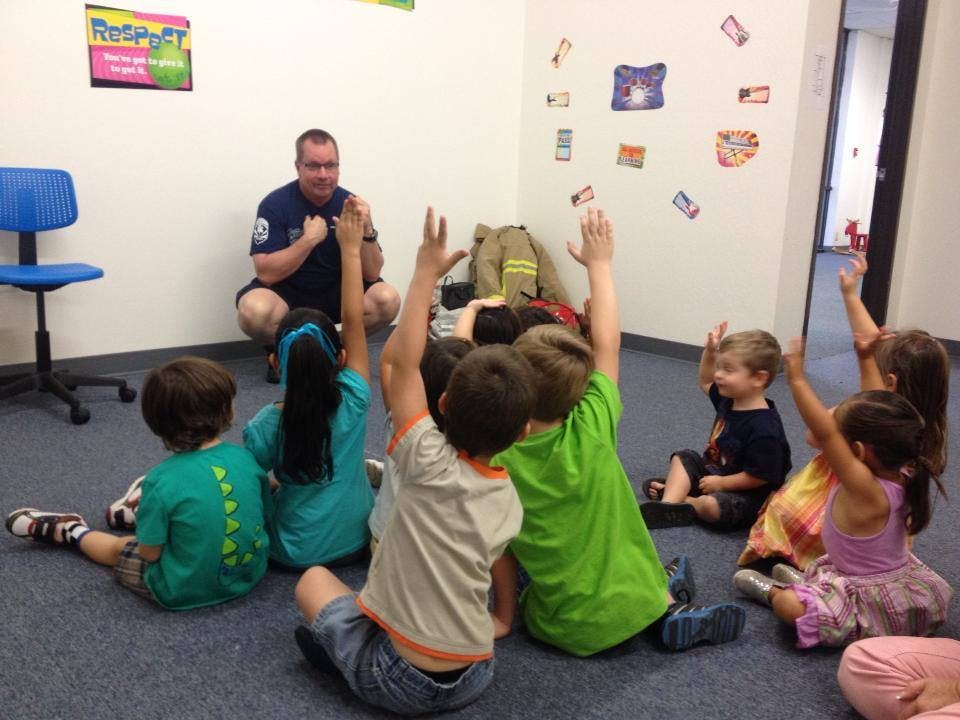 Adult speaking to a group of children with raised hands in a classroom