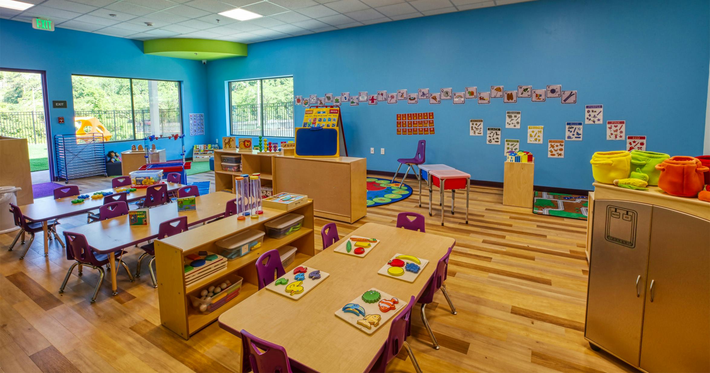 Classroom with tables, chairs, learning materials, and playground visible outside