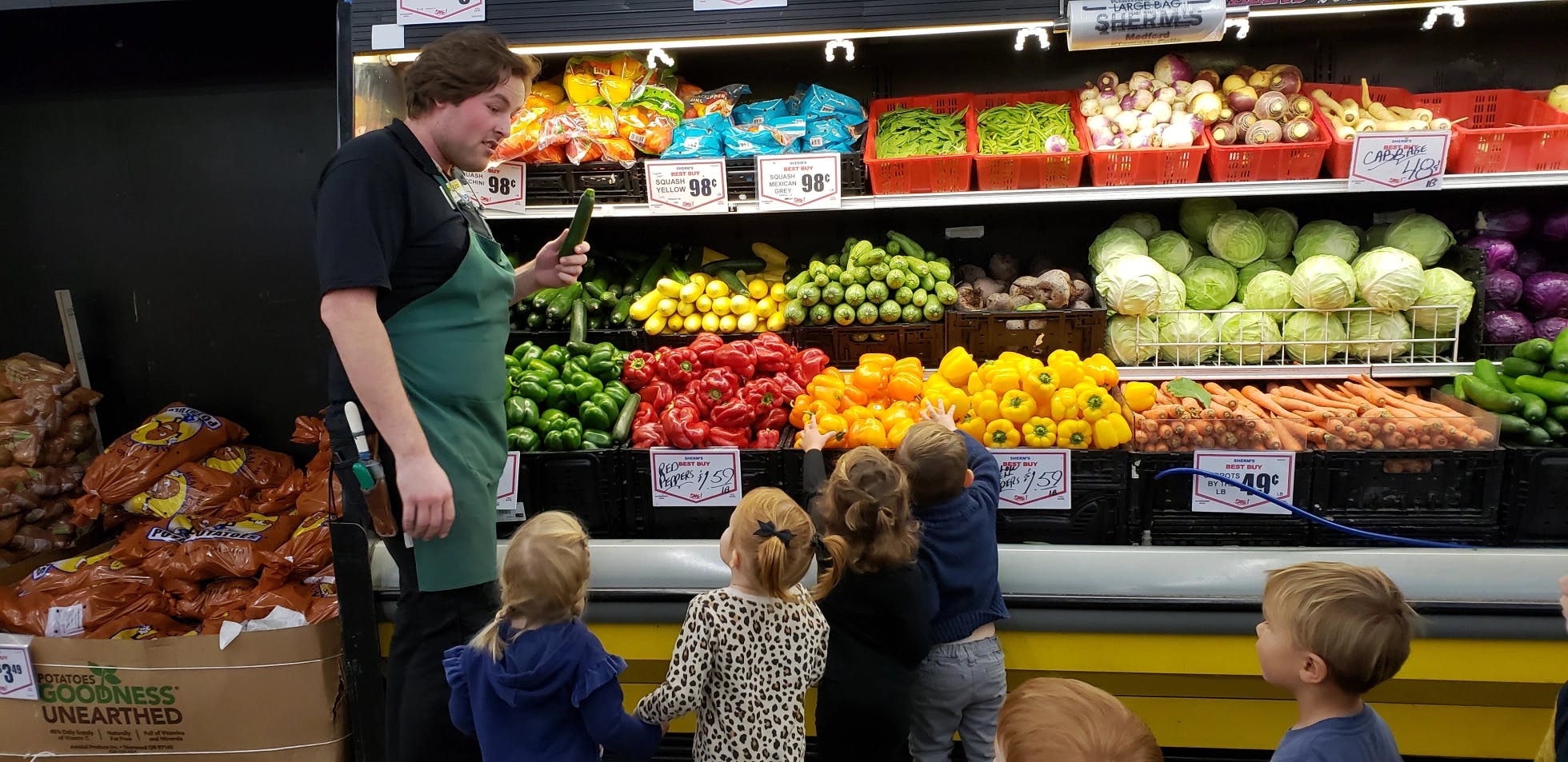 Person in apron talking to children in front of produce display