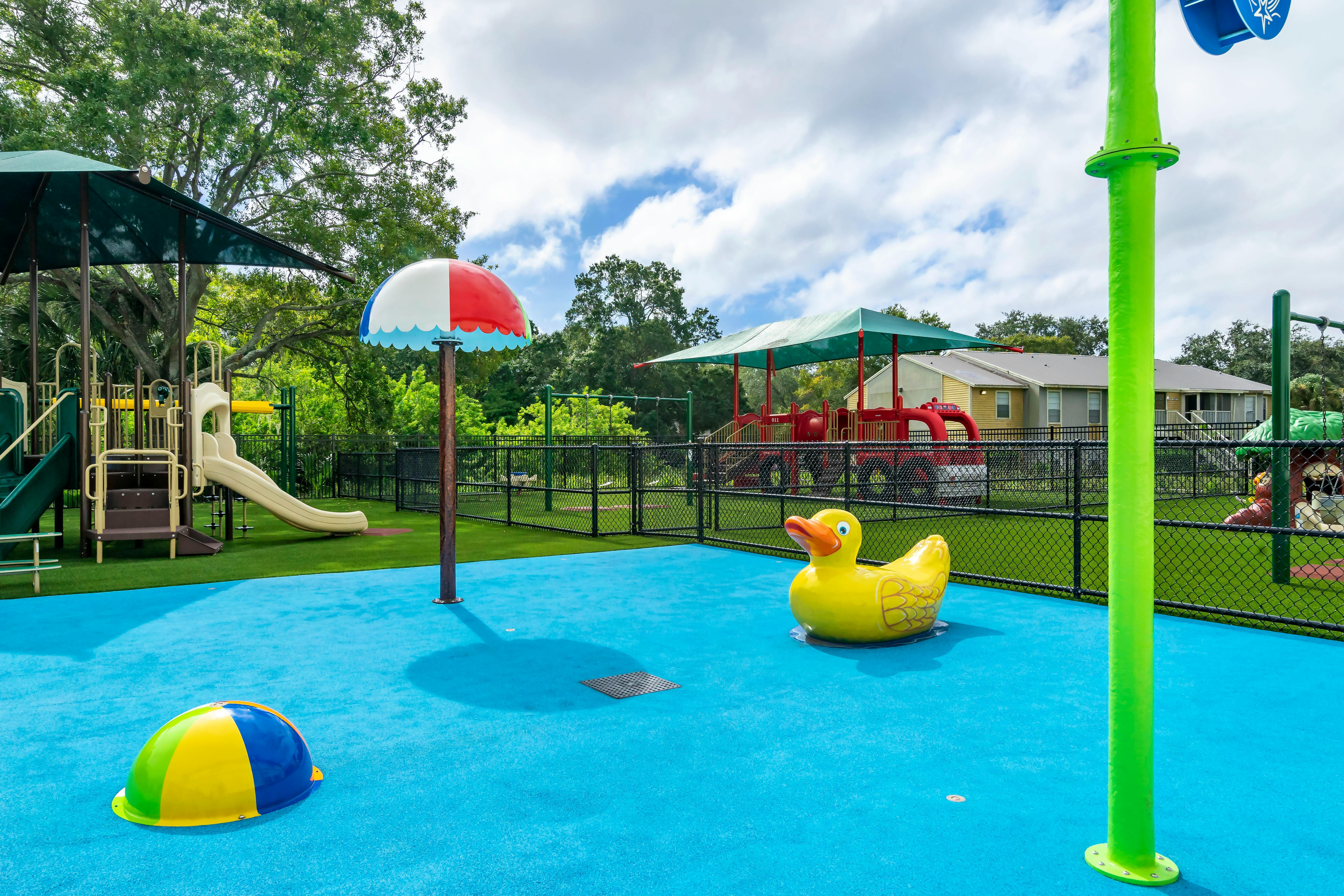 A spray ground with a yellow duck and colorful umbrella sprayer, playground in background