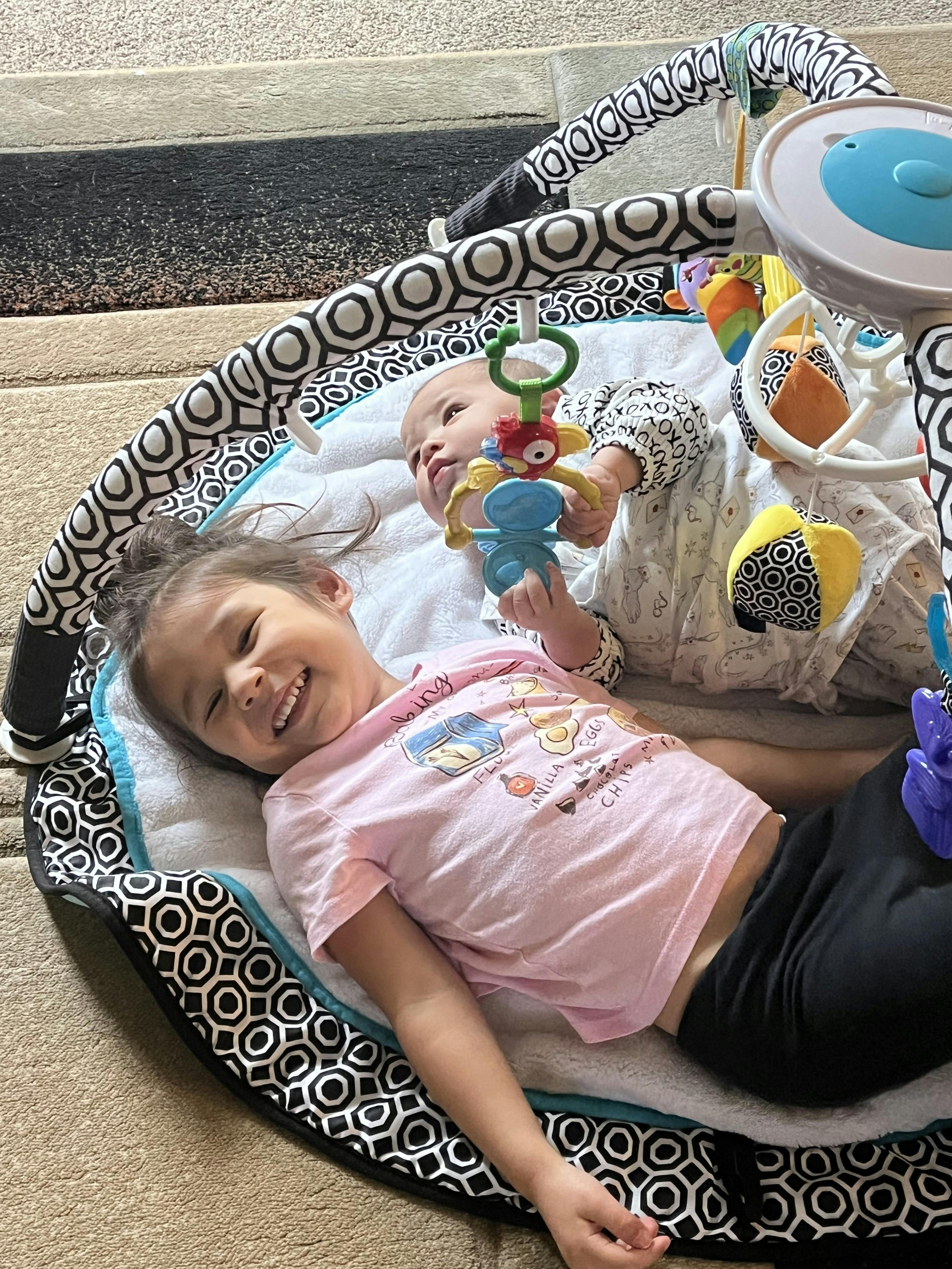 A child and an infant lying on a play mat with hanging toys