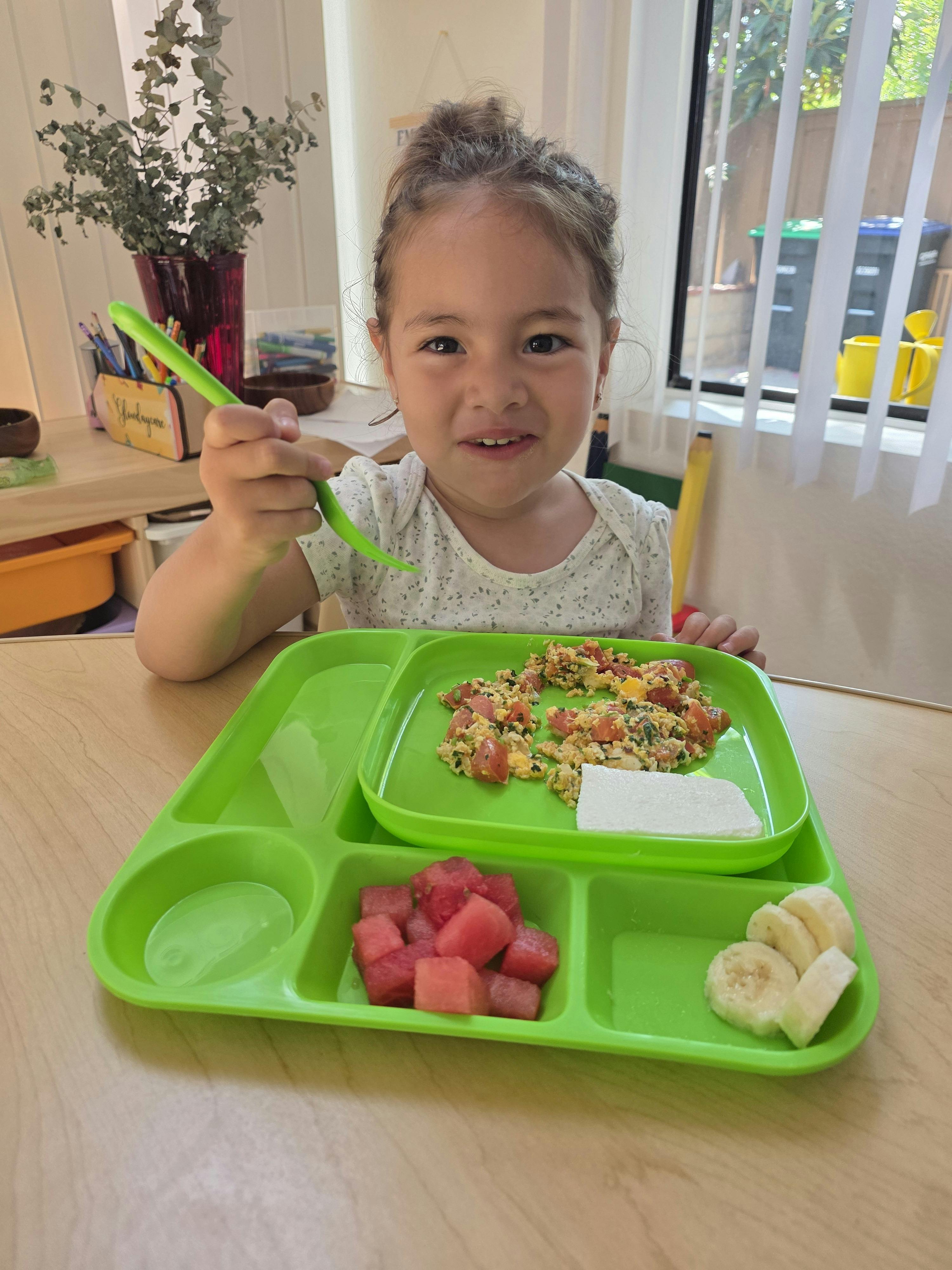 Child holding a spoon while sitting at a table with a meal on a divided tray