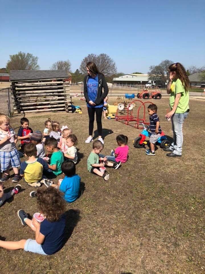 Two adults supervising a group of children sitting and playing on the ground outdoors