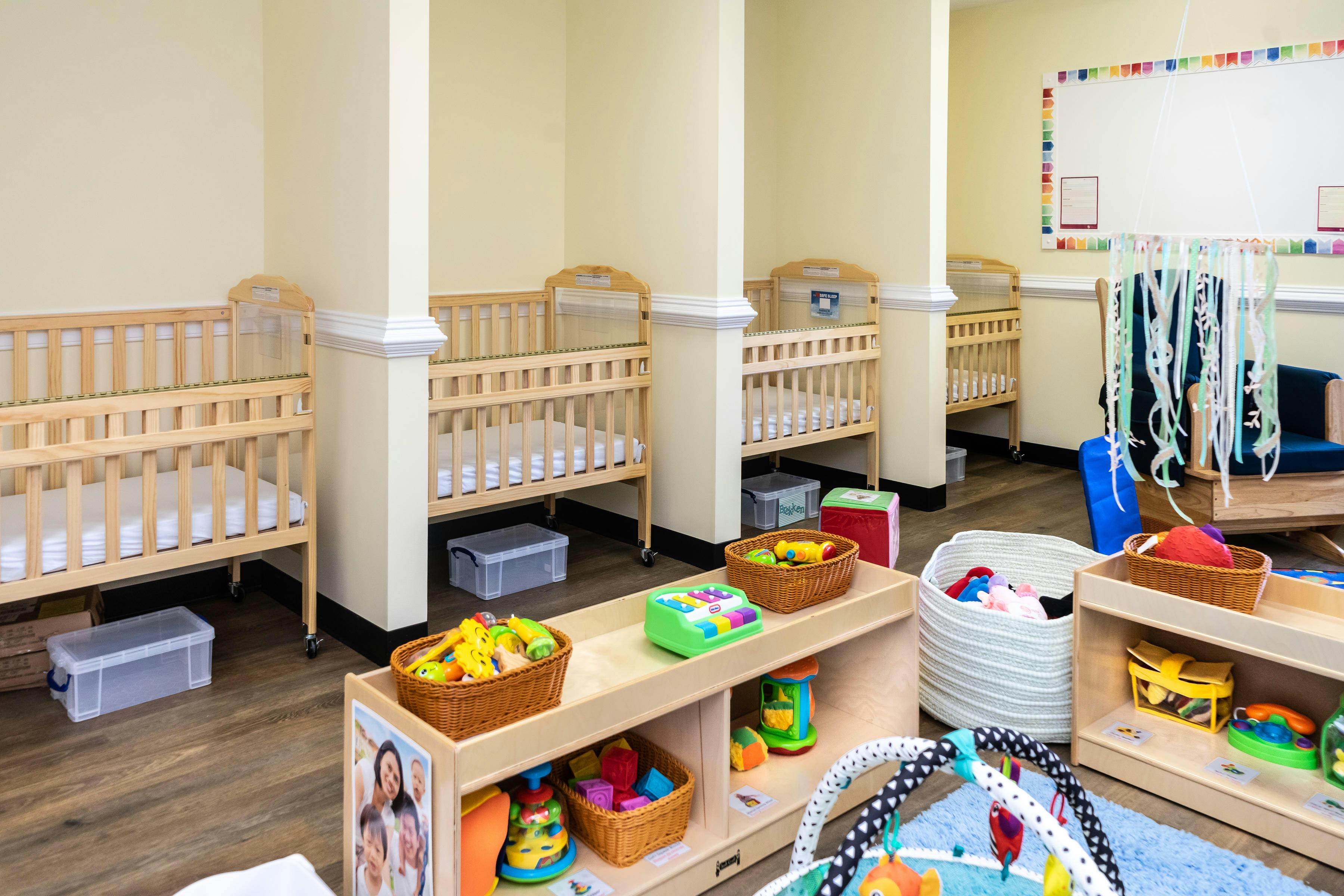 multiple wooden cribs lined against a light yellow wall, with infant play items in the foreground