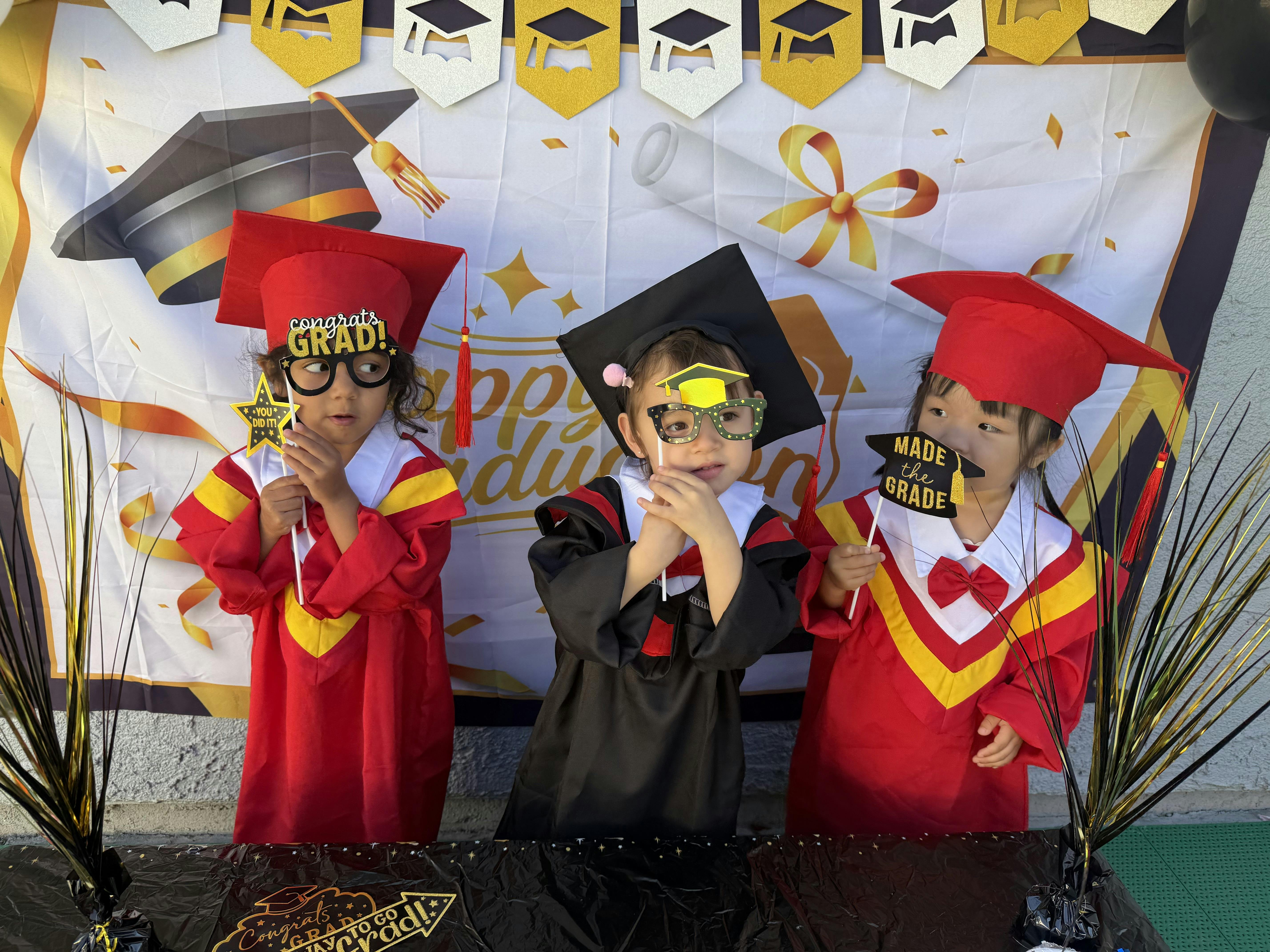 Three children in graduation caps and gowns holding props against a banner