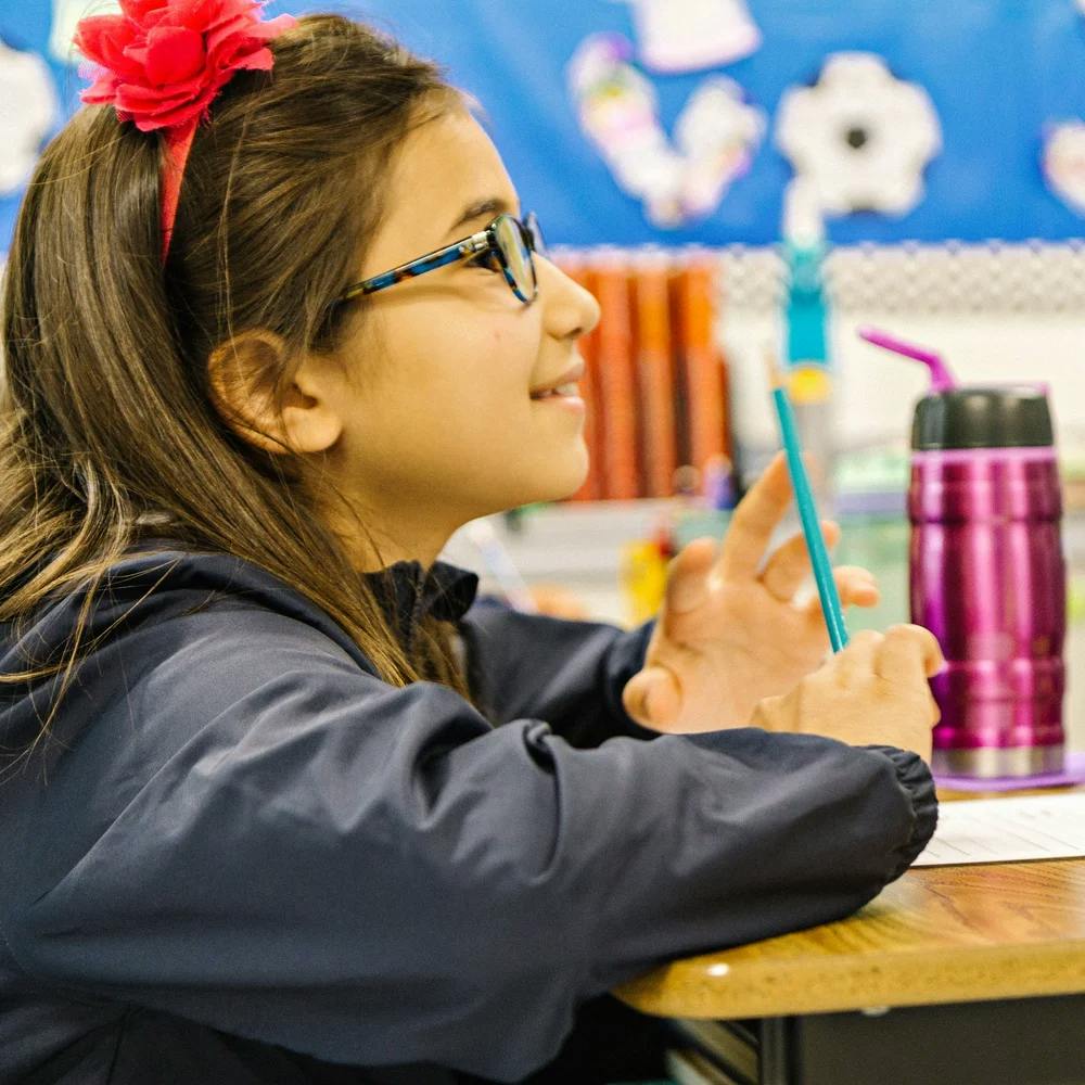 Child wearing glasses and a red headband, holding a pencil in a classroom