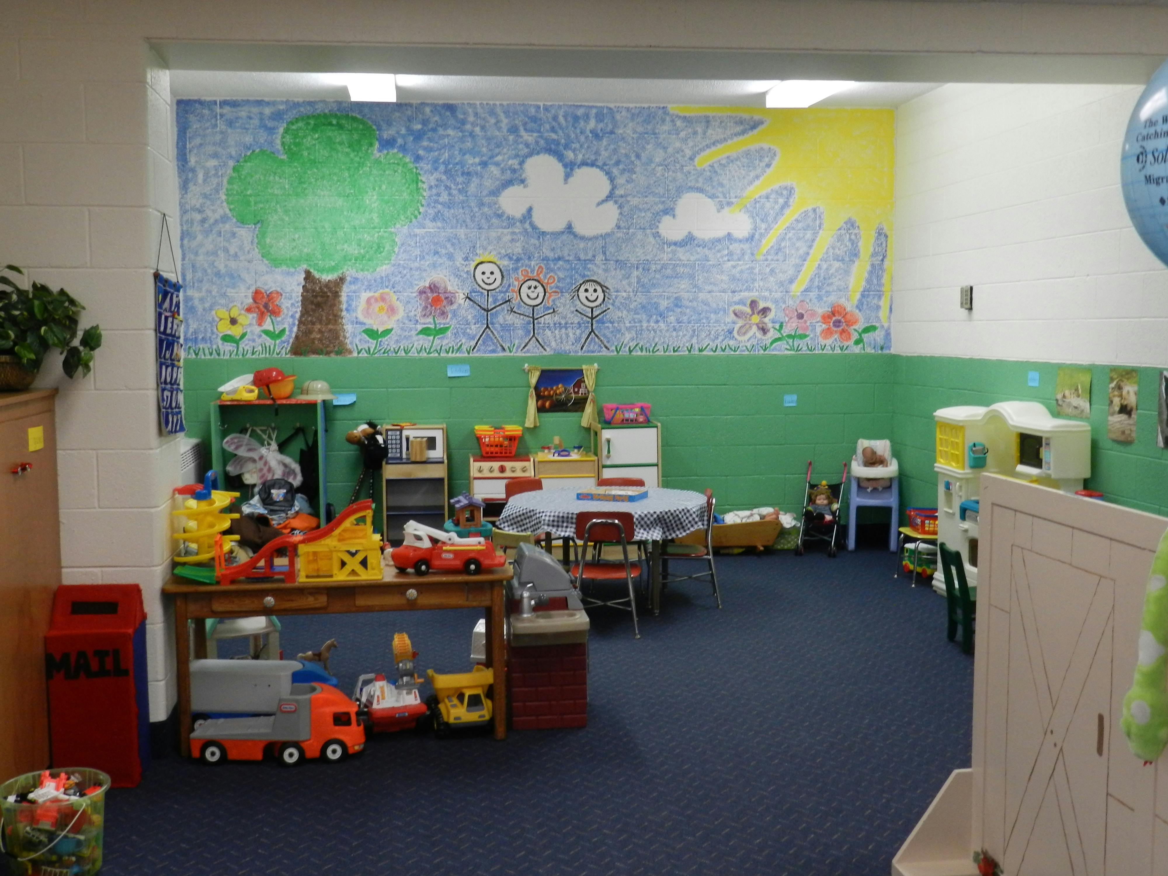 Playroom with toys, tables, and a mural of a tree, sun, and clouds