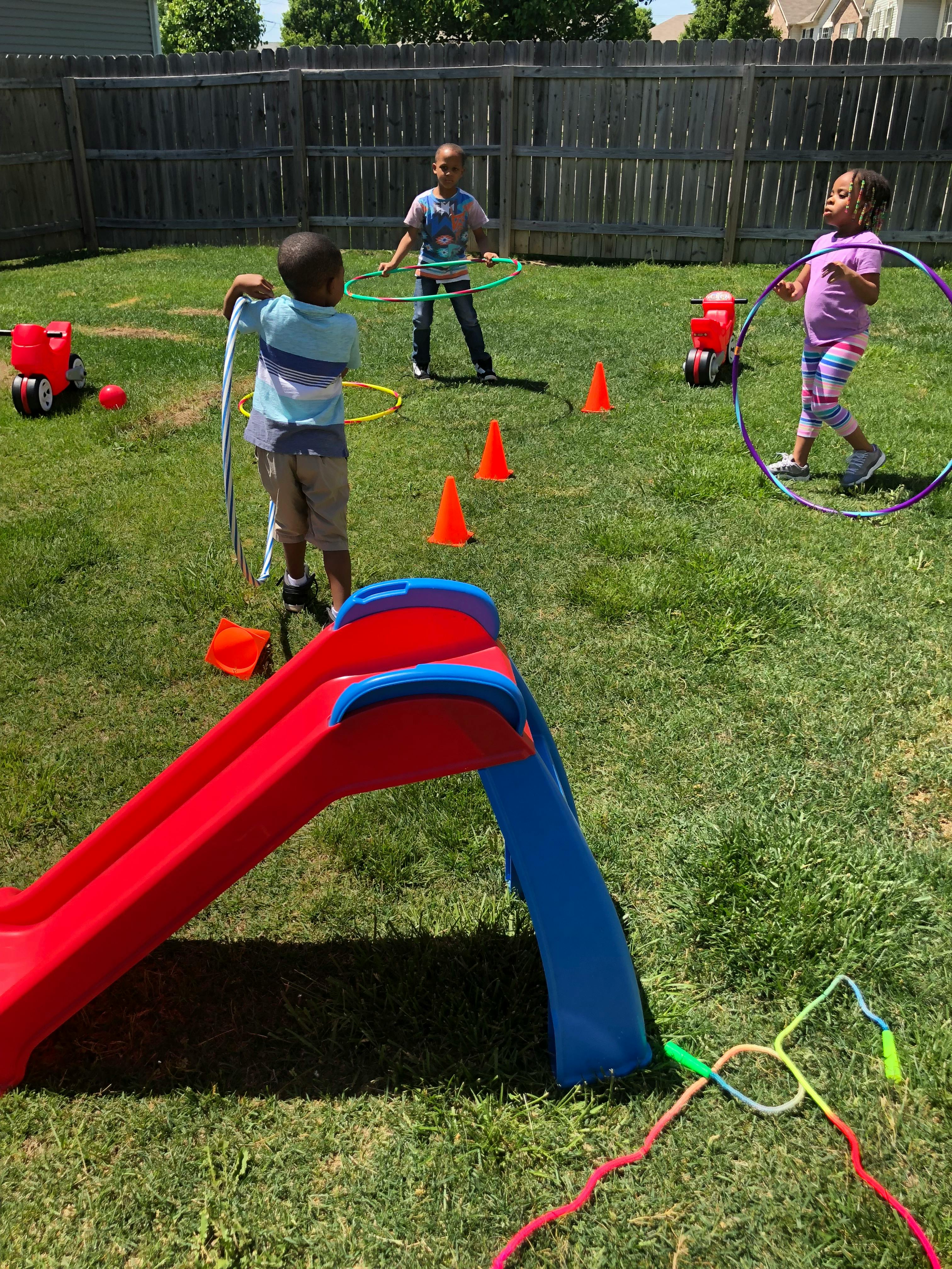 Three children playing with hula hoops on a grass lawn near a slide