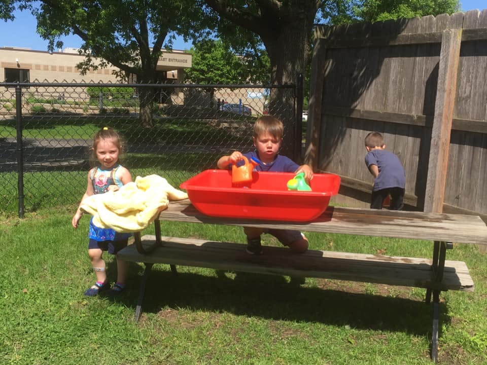 Three children playing outdoors with a red water table on a picnic table