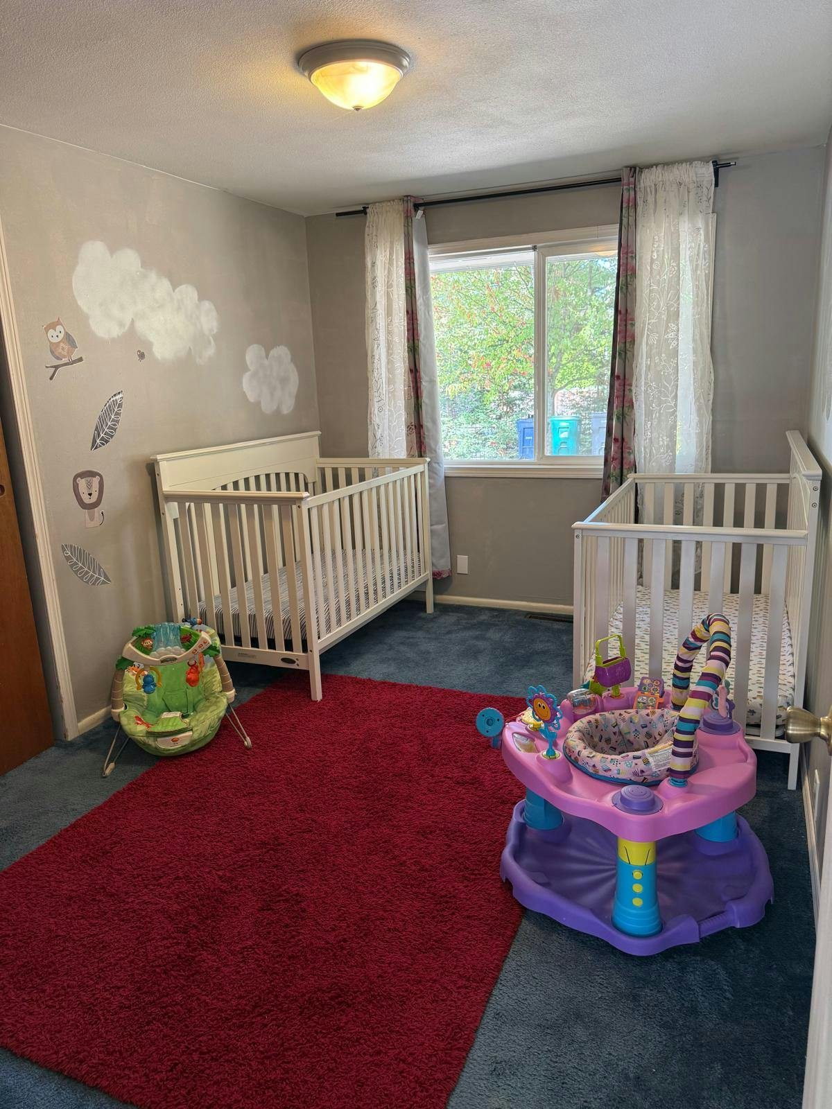 Two cribs and baby activity centers in a room with a red rug