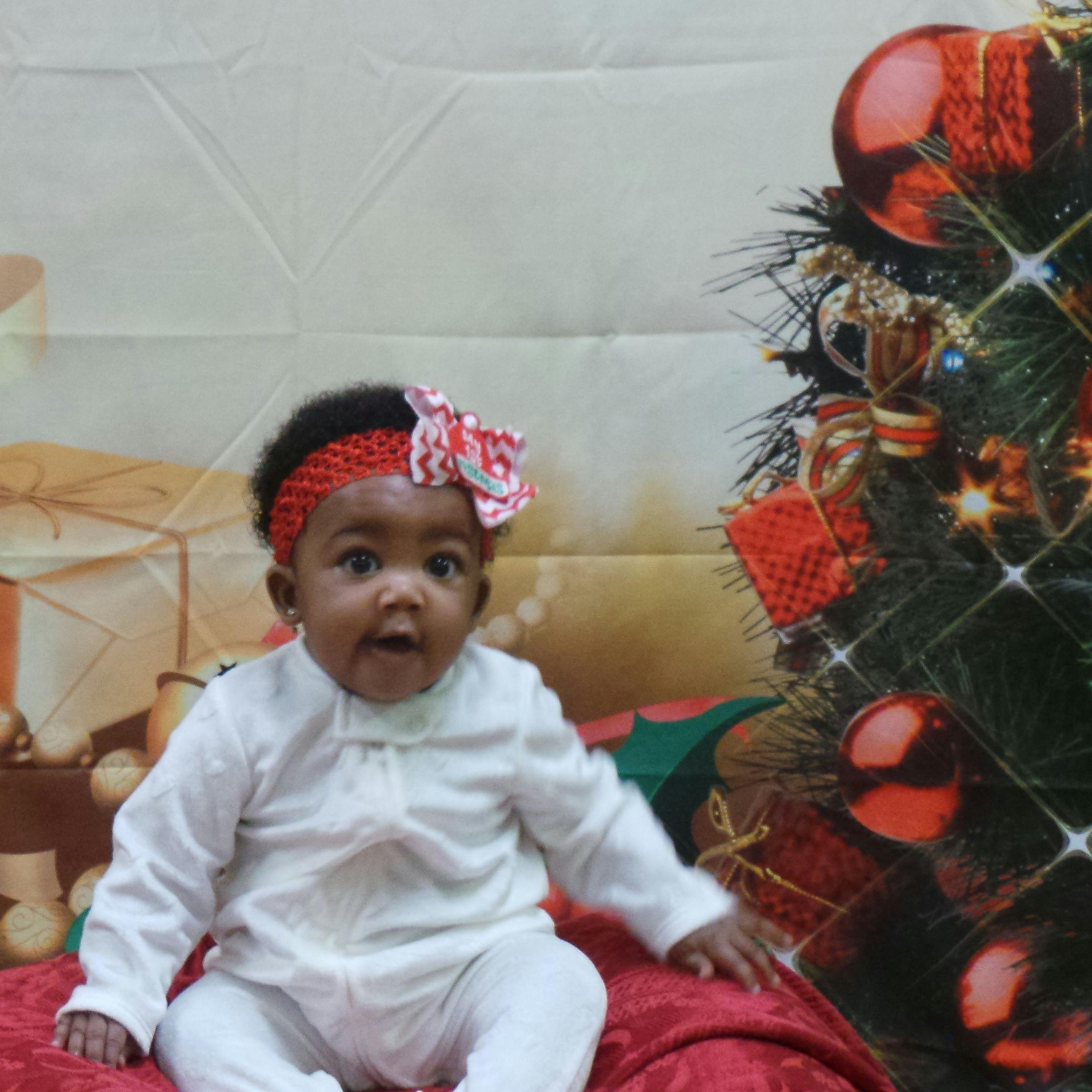 Child wearing a red headband with a bow, sitting next to a Christmas tree