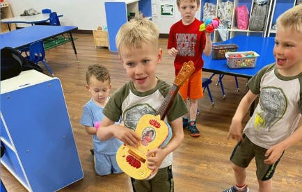 Child holding a toy guitar with other children in a room