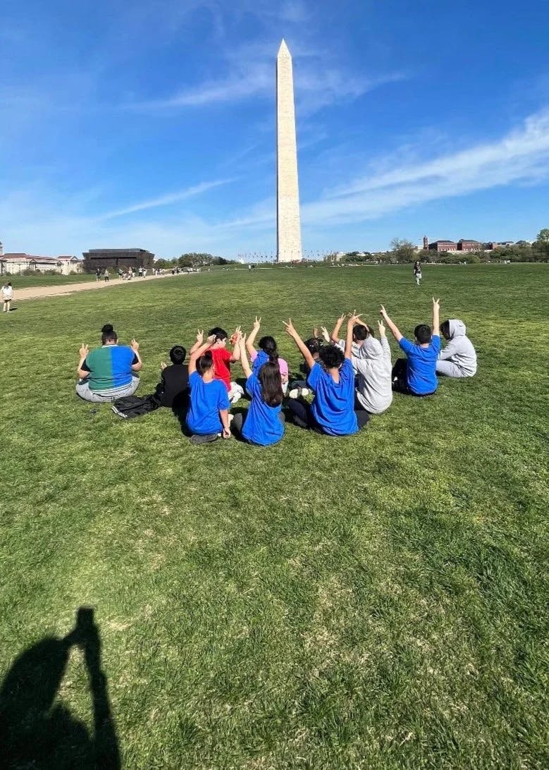Group of children seated on grass, making peace signs at a tall monument