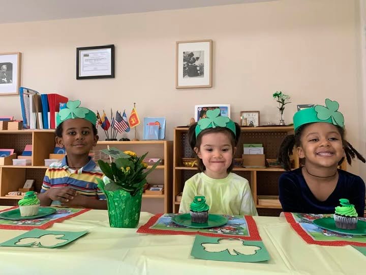 Three children wearing green shamrock headbands sitting at a table with cupcakes